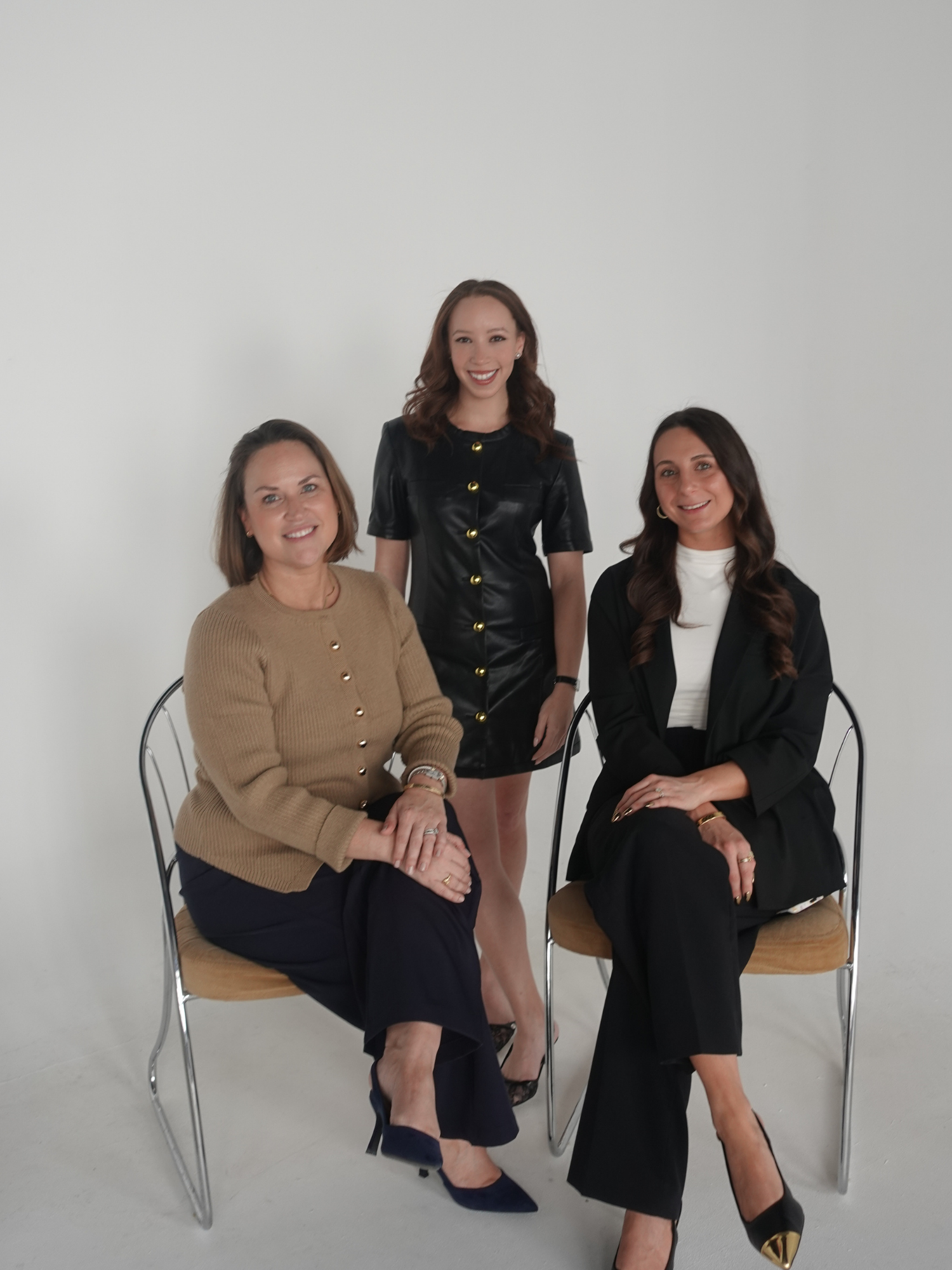 Three women in professional attire posing in front of a plain white wall, two sitting on chairs and one standing in the middle.