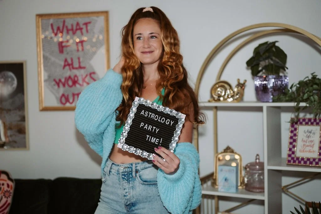 A woman with long red hair, wearing a blue fuzzy cardigan and a green top, holding a black letter board that reads "Astrology Party Time!" inside a decorated room.