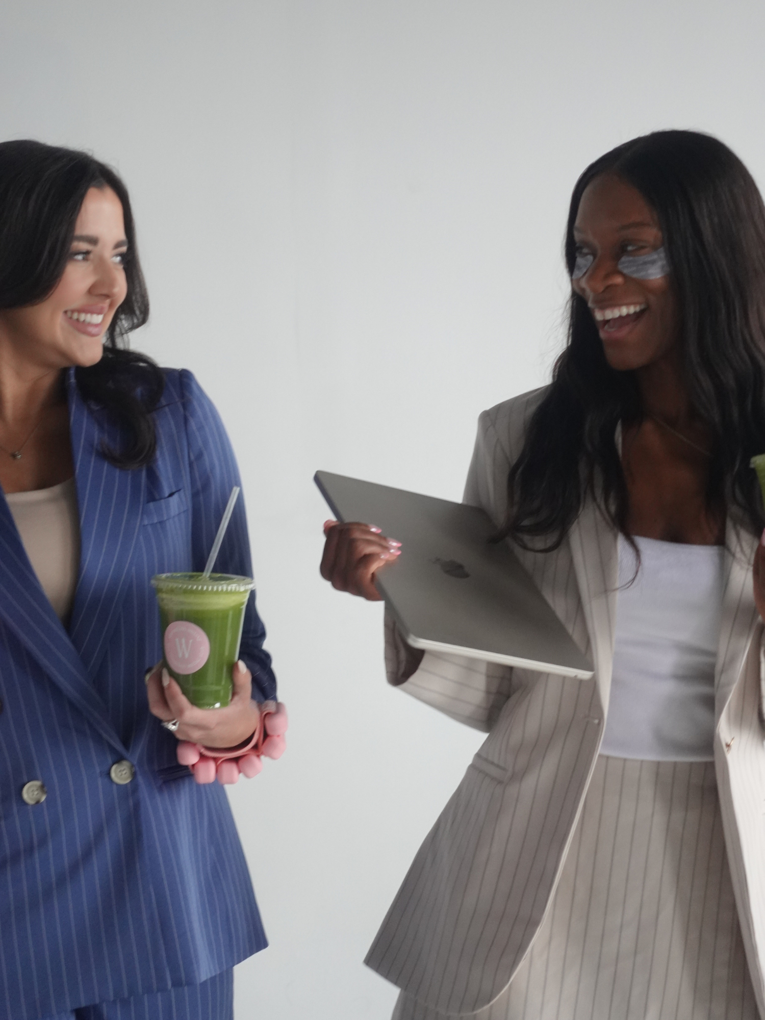 Two women in professional attire smiling and talking, one holding a green smoothie with a straw, and the other holding a gray tablet.