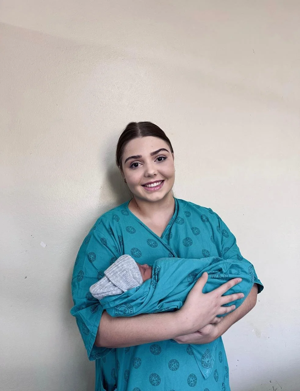 A smiling young woman in a blue hospital gown holding a newborn baby wrapped in a gray blanket.