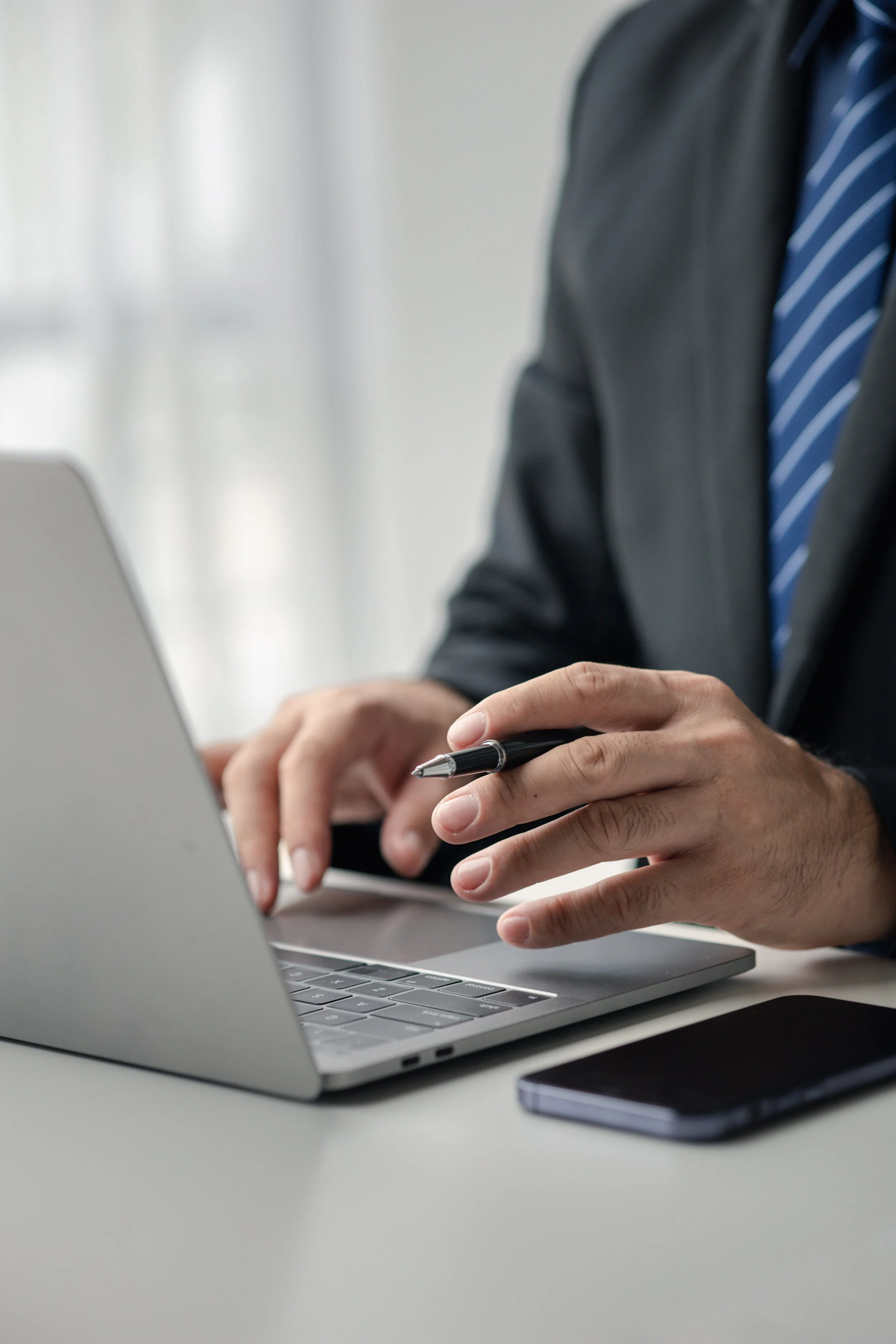 Close-up of a person wearing a business suit with a striped blue tie, typing on a laptop keyboard, holding a pen in one hand, with a smartphone on the table nearby.
