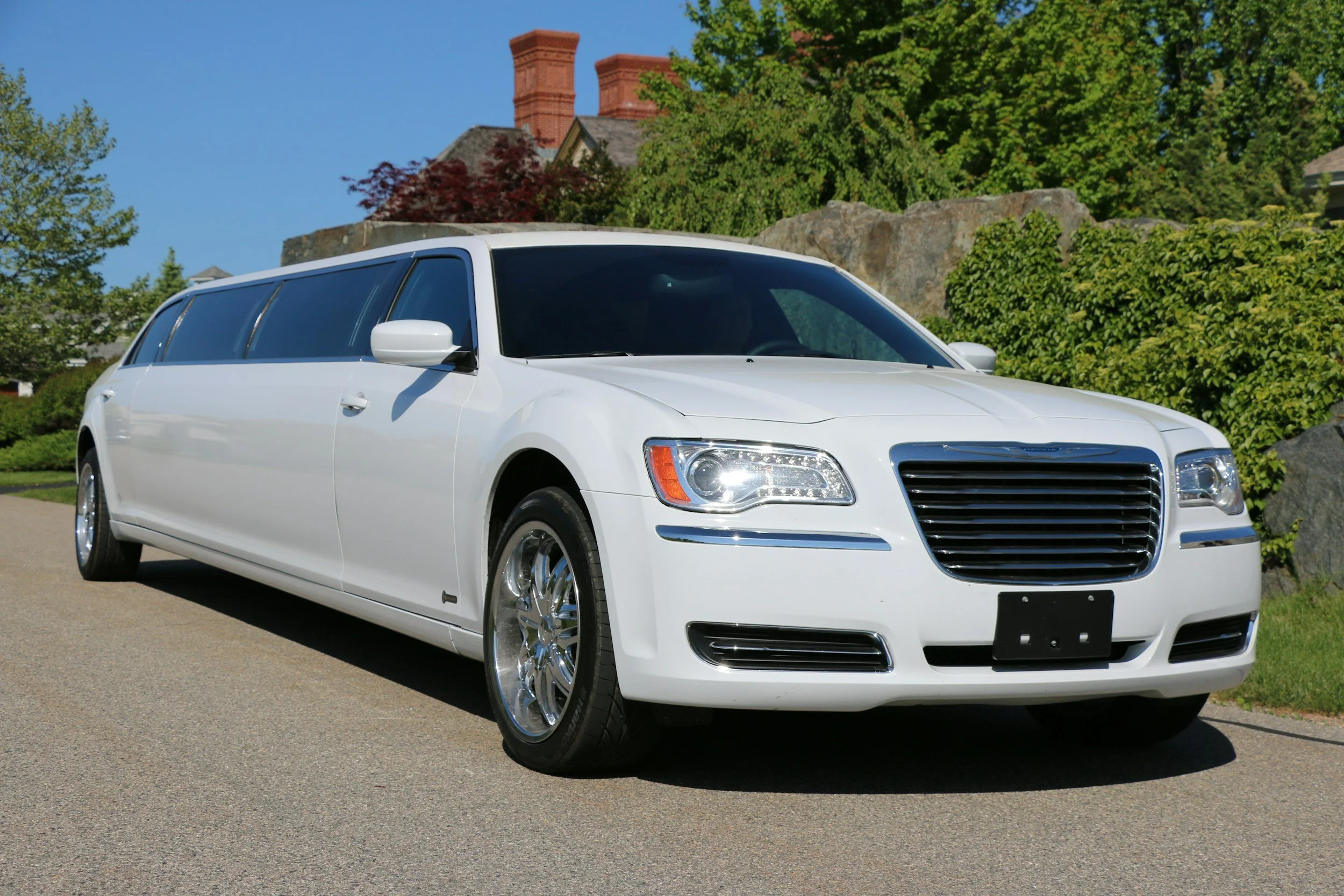 White limousine is parked on a paved road with green trees and a house with chimneys in the background on a sunny day.