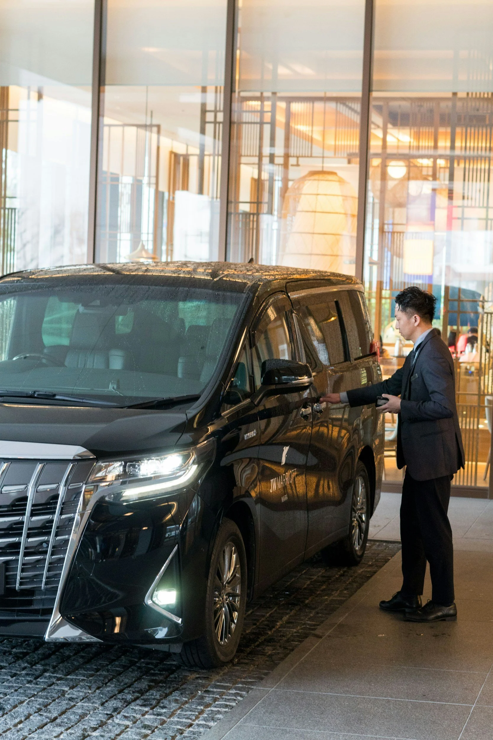 A chauffeur in the livery industry wearing business attire unlocks a black luxury SUV parked outside a modern building with large glass windows and wooden accents.