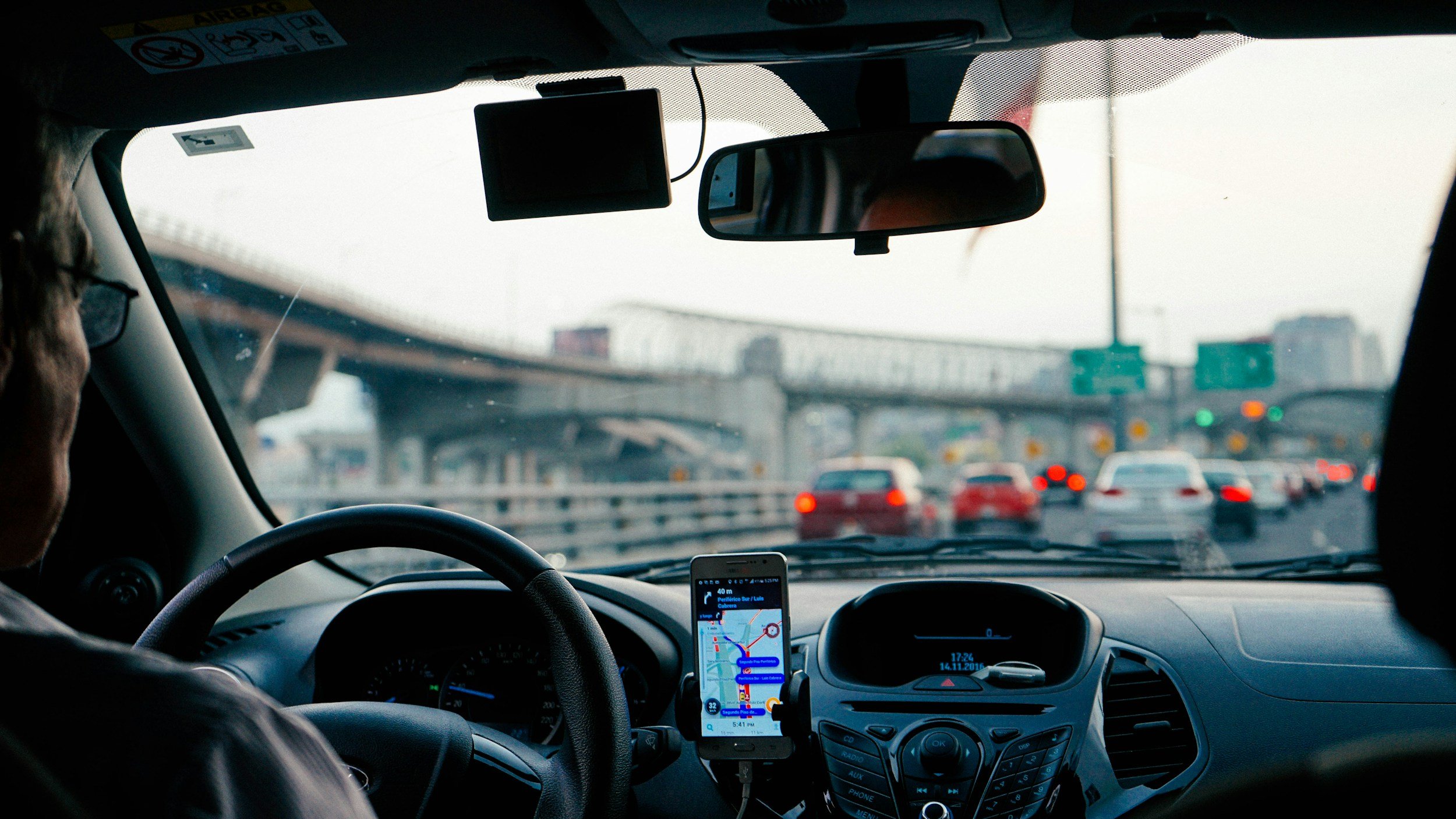 View from inside a car on a highway during daytime, showing the dashboard, steering wheel, GPS on the windshield, and other vehicles ahead, with an overpass bridge in the distance.