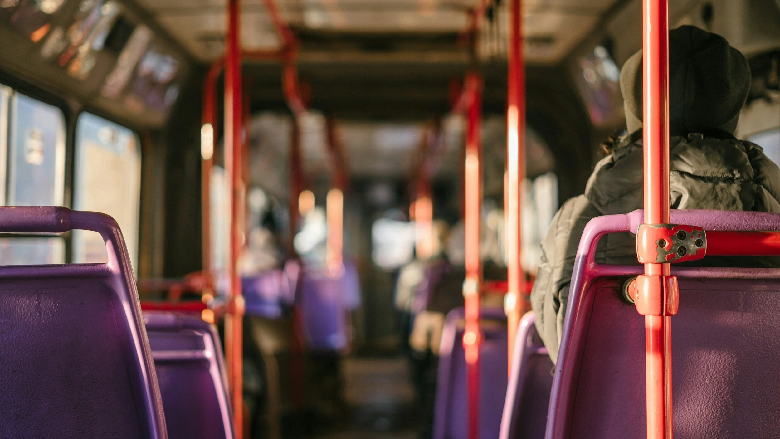 Inside a bus with empty purple seats and an orange pole, with a person sitting on the right side wearing a gray hoodie and a dark coat.
