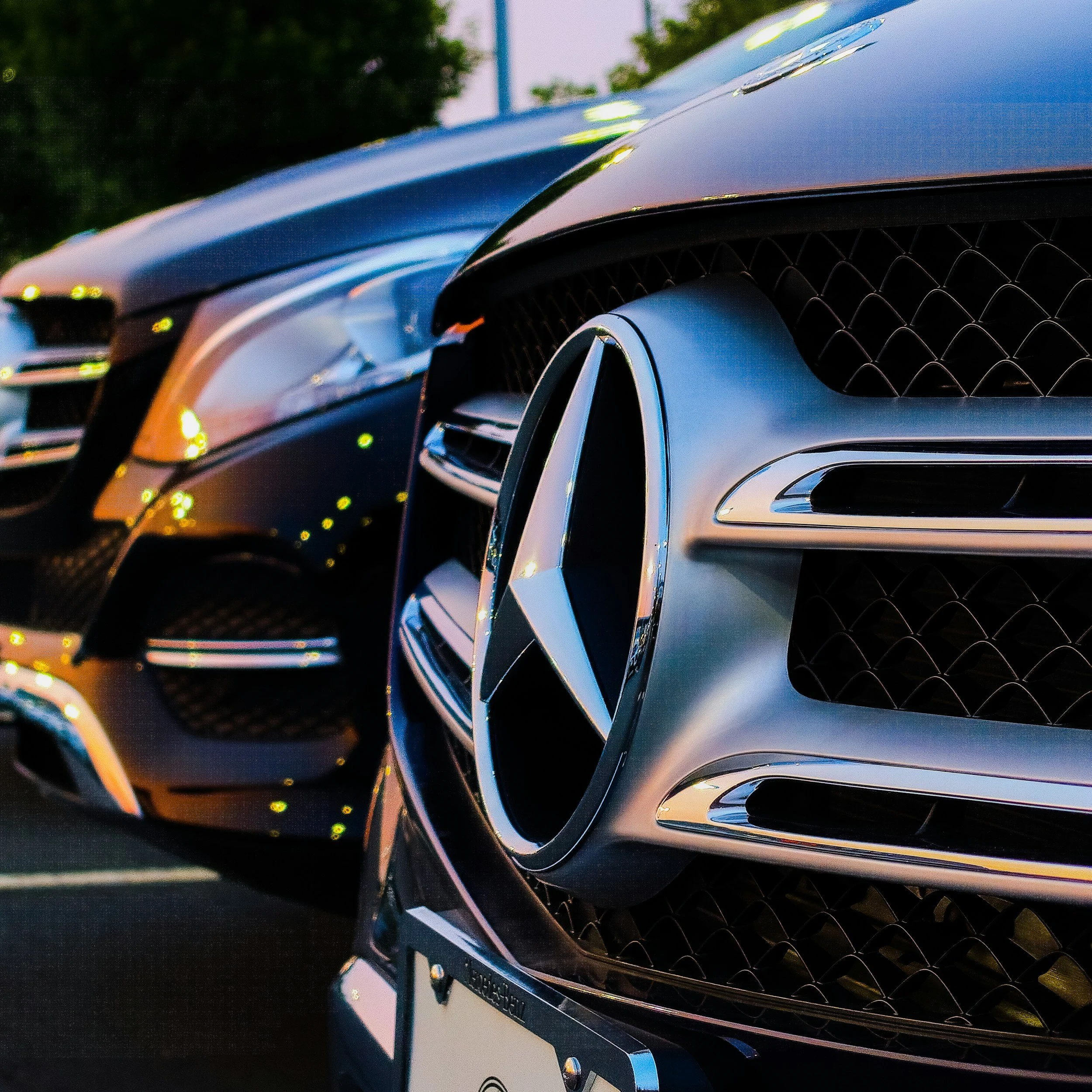 Close-up of the front end of a black luxury Mercedes-Benz car, showing the grille, emblem, and part of the headlight with a blurred background of green trees.