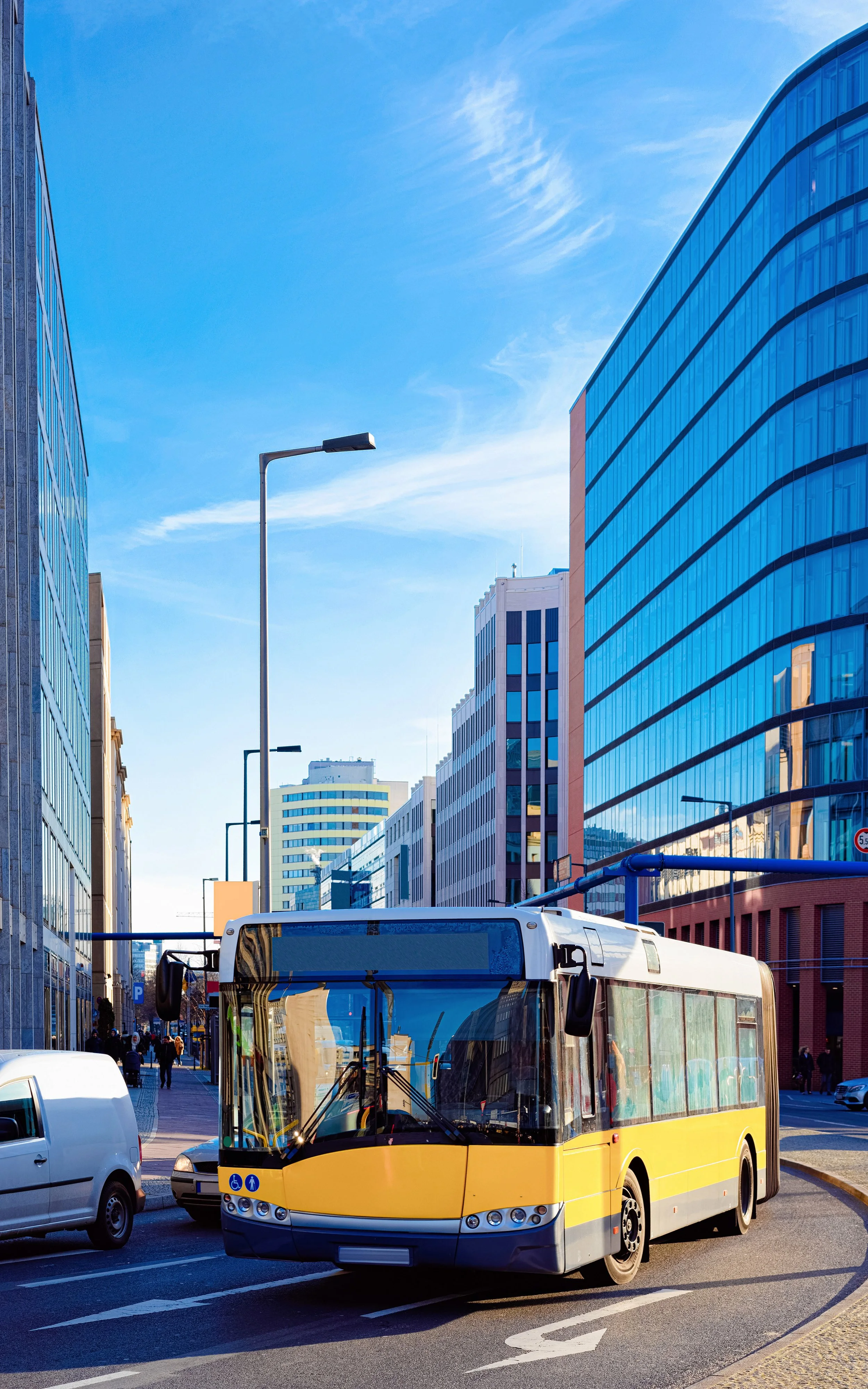 Yellow city bus on a city street with tall modern glass and concrete buildings and a blue sky with clouds in the background, daytime.