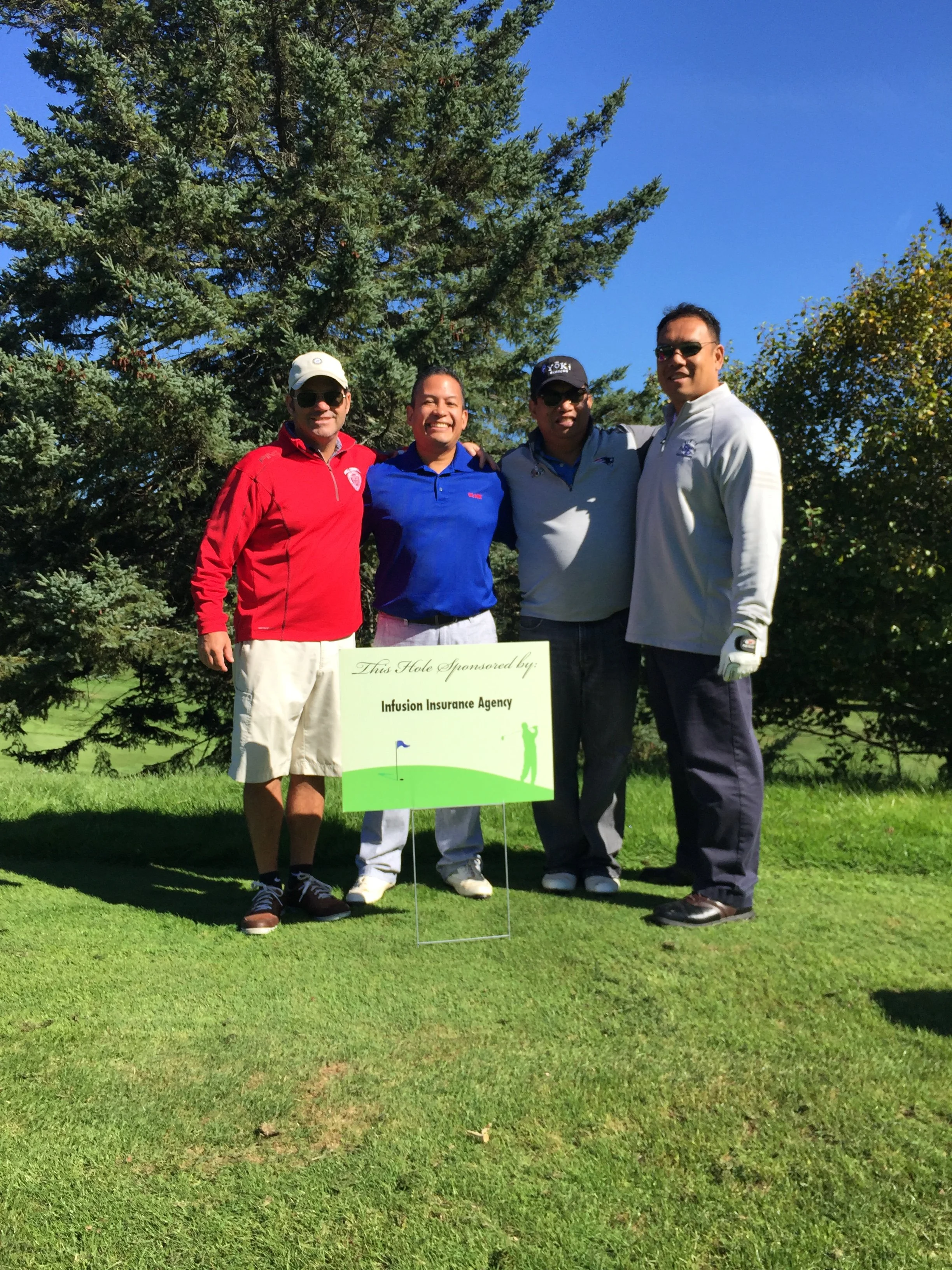 Four men, including Art Chapang, are standing on a golf course, smiling and posing for the photo. They are near a sign that reads "This Hole Sponsored by: Infusion Insurance Agency." There are trees in the background and the sky is clear and blue.