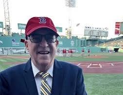 Bob Crescenzo, wearing glasses, a red Boston Red Sox cap, a dark suit, and a striped tie, is standing in a baseball stadium near the field.