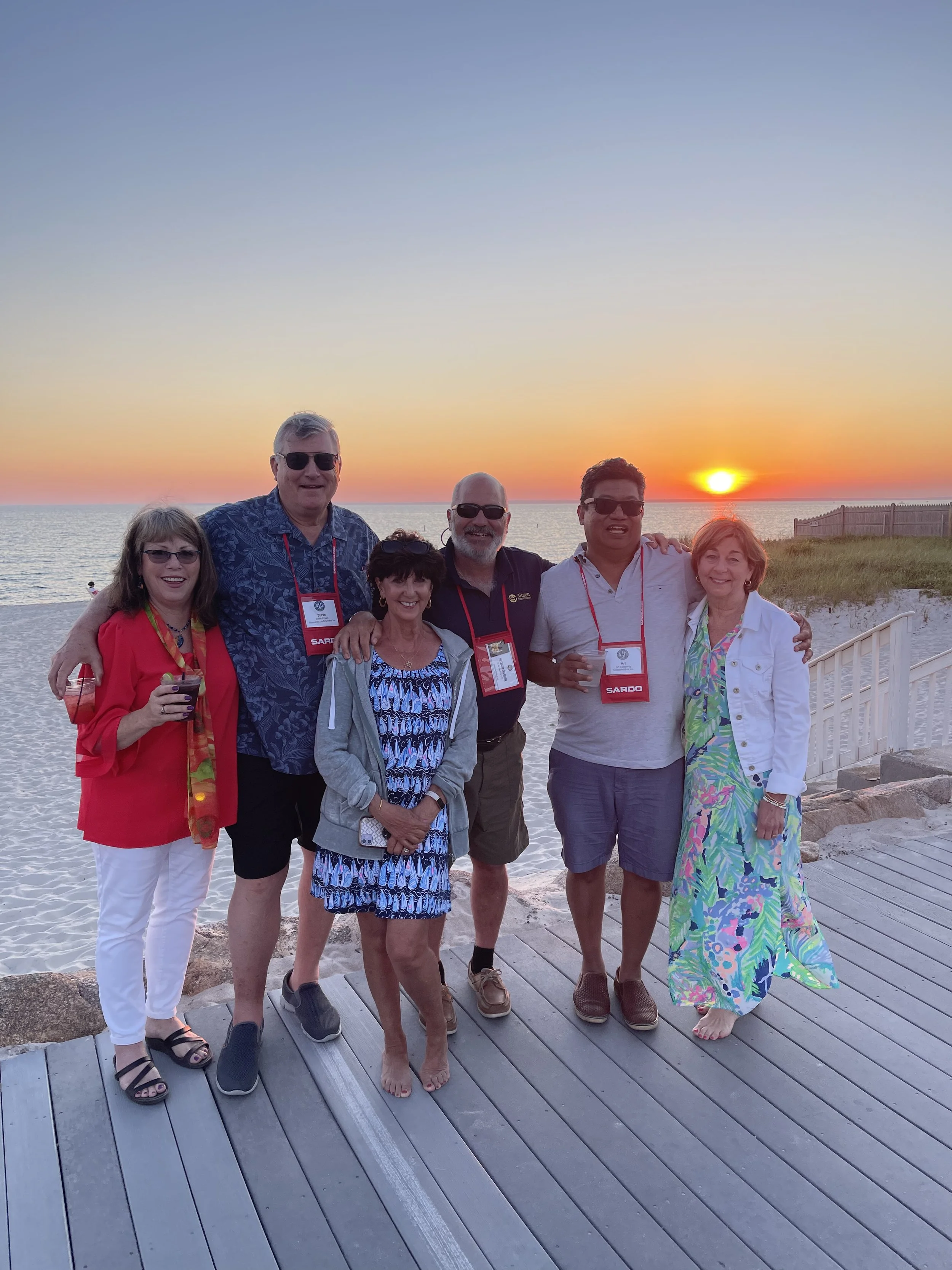 Infusion Insurance team members, including founder Art Catapang, standing on a wooden boardwalk near the beach at sunset, with the ocean and a fence in the background.