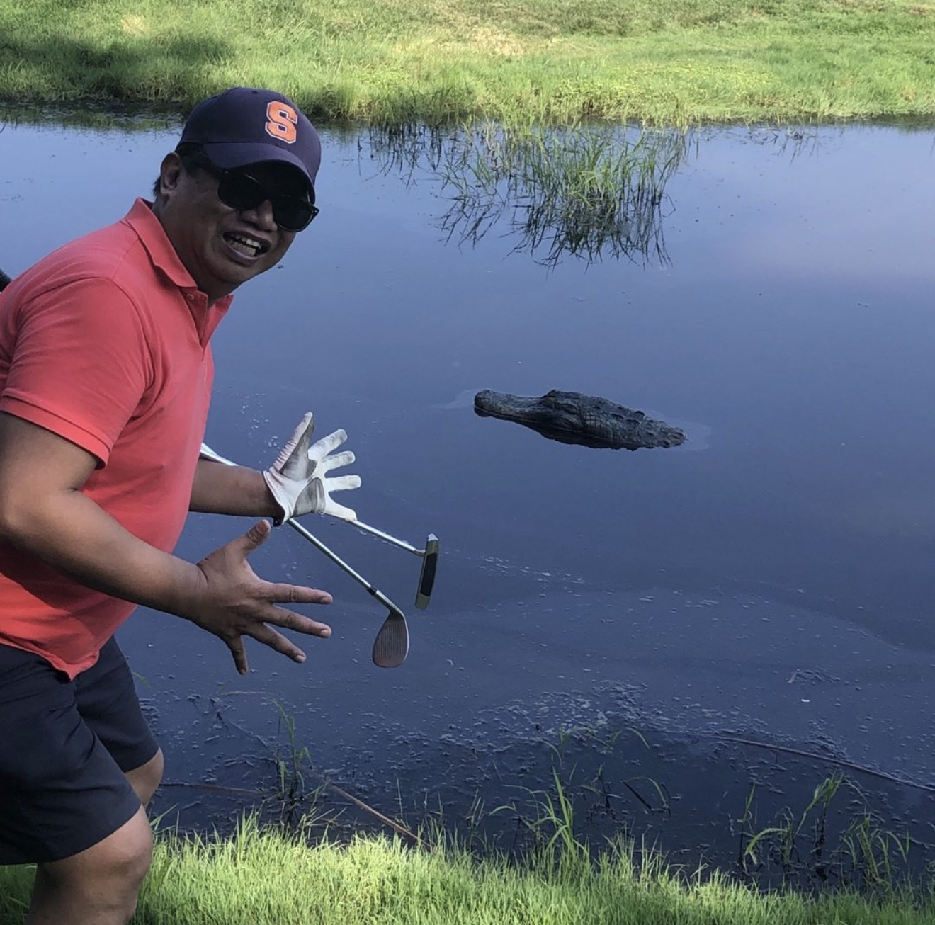 Art Catapang in a red polo shirt, sunglasses, and a baseball cap, catching a small alligator in a pond with a golf club, with green grass and water in the background.