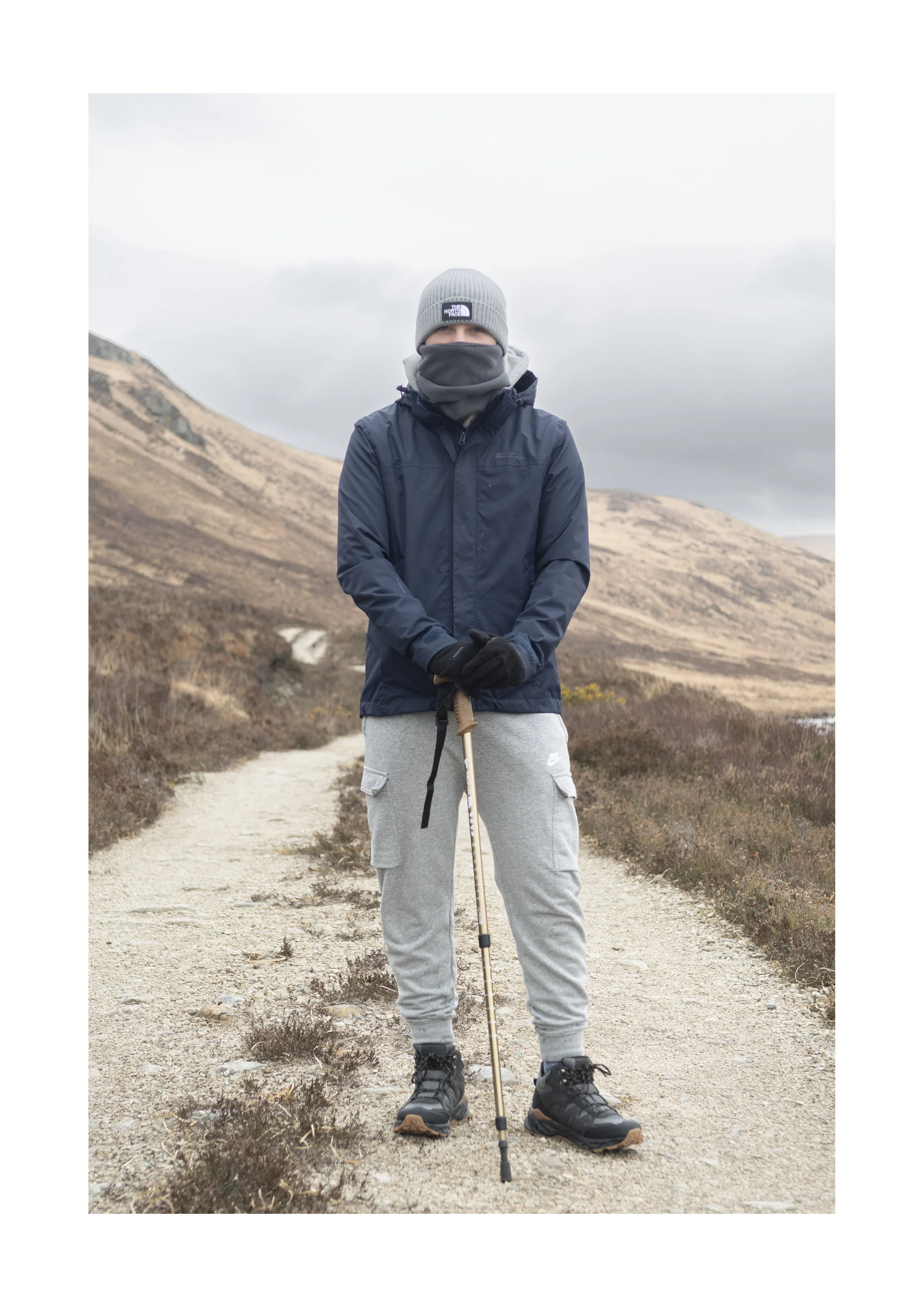Person standing outdoors on a dirt trail, wearing a gray beanie, a dark jacket, gray sweatpants, and hiking boots, holding a hiking pole, with mountains and cloudy sky in the background.