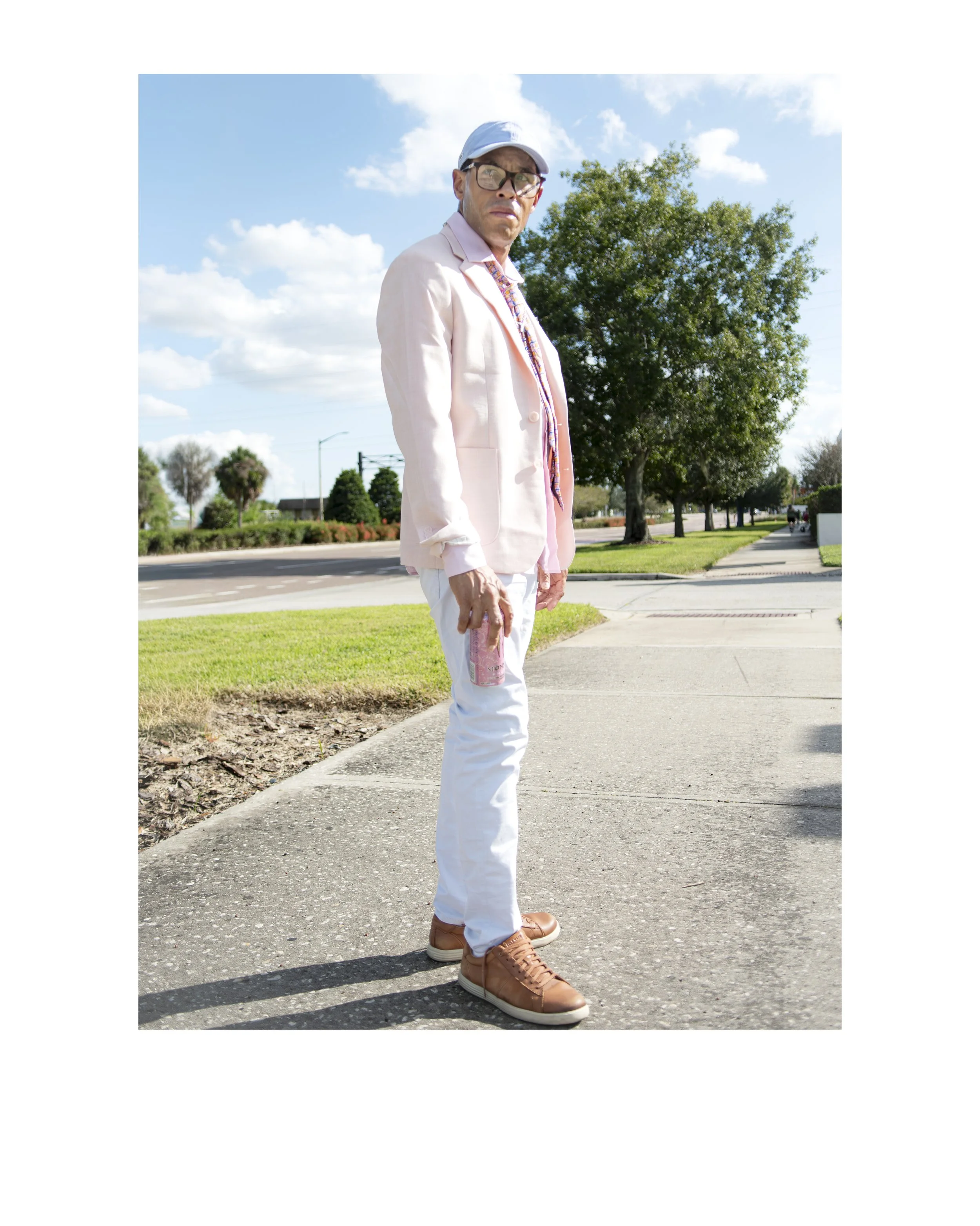 Man in sunglasses, light pink blazer, and white pants standing on sidewalk with trees and blue sky in background.