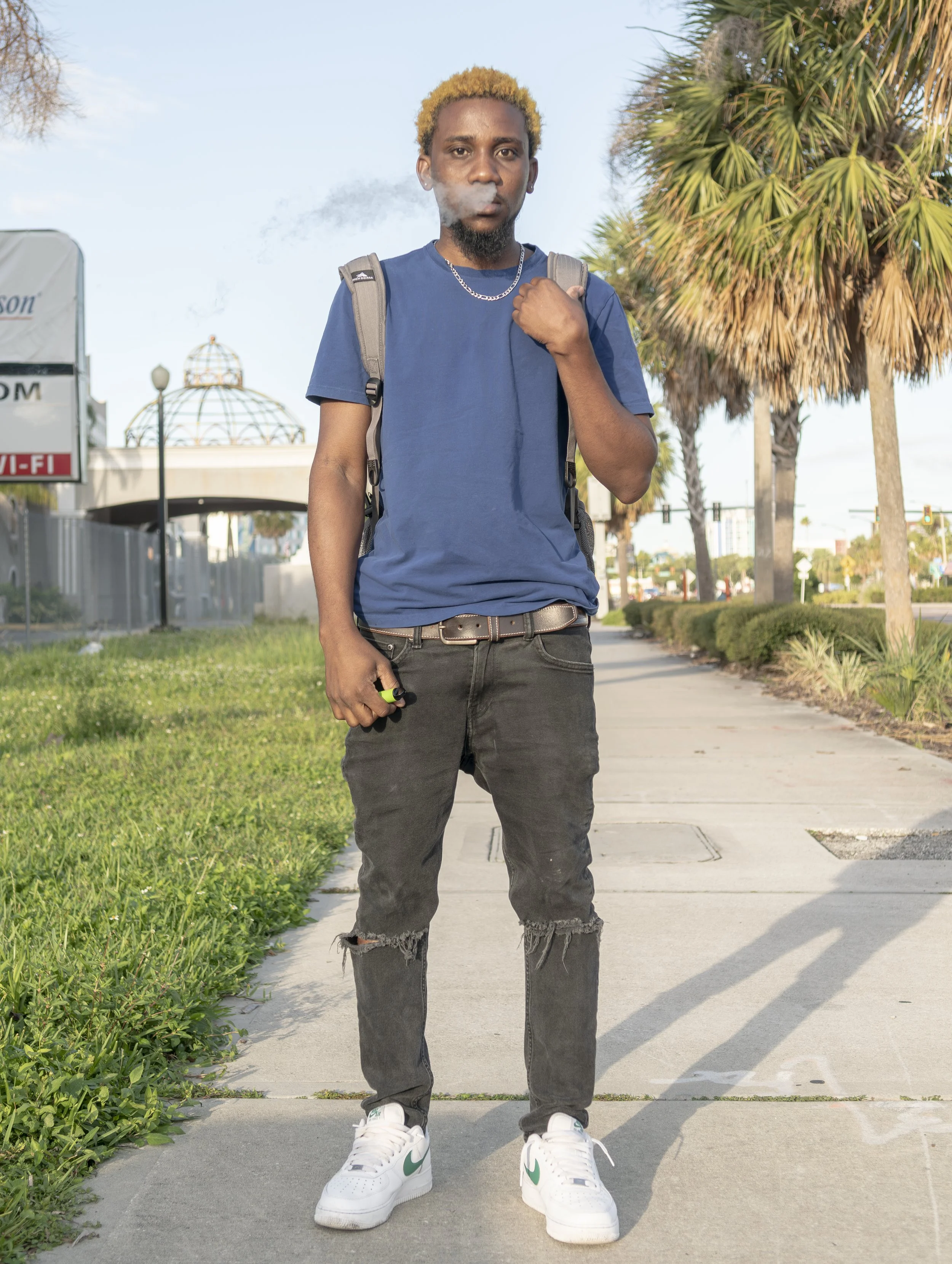 A young man with blond hair and a goatee standing outdoors on a sidewalk, wearing a blue t-shirt, black ripped jeans, white sneakers with green accents, a gray backpack, a silver chain necklace, and holding a remote control in his right hand. He is blowing smoke from his mouth, with trees and a building visible in the background.