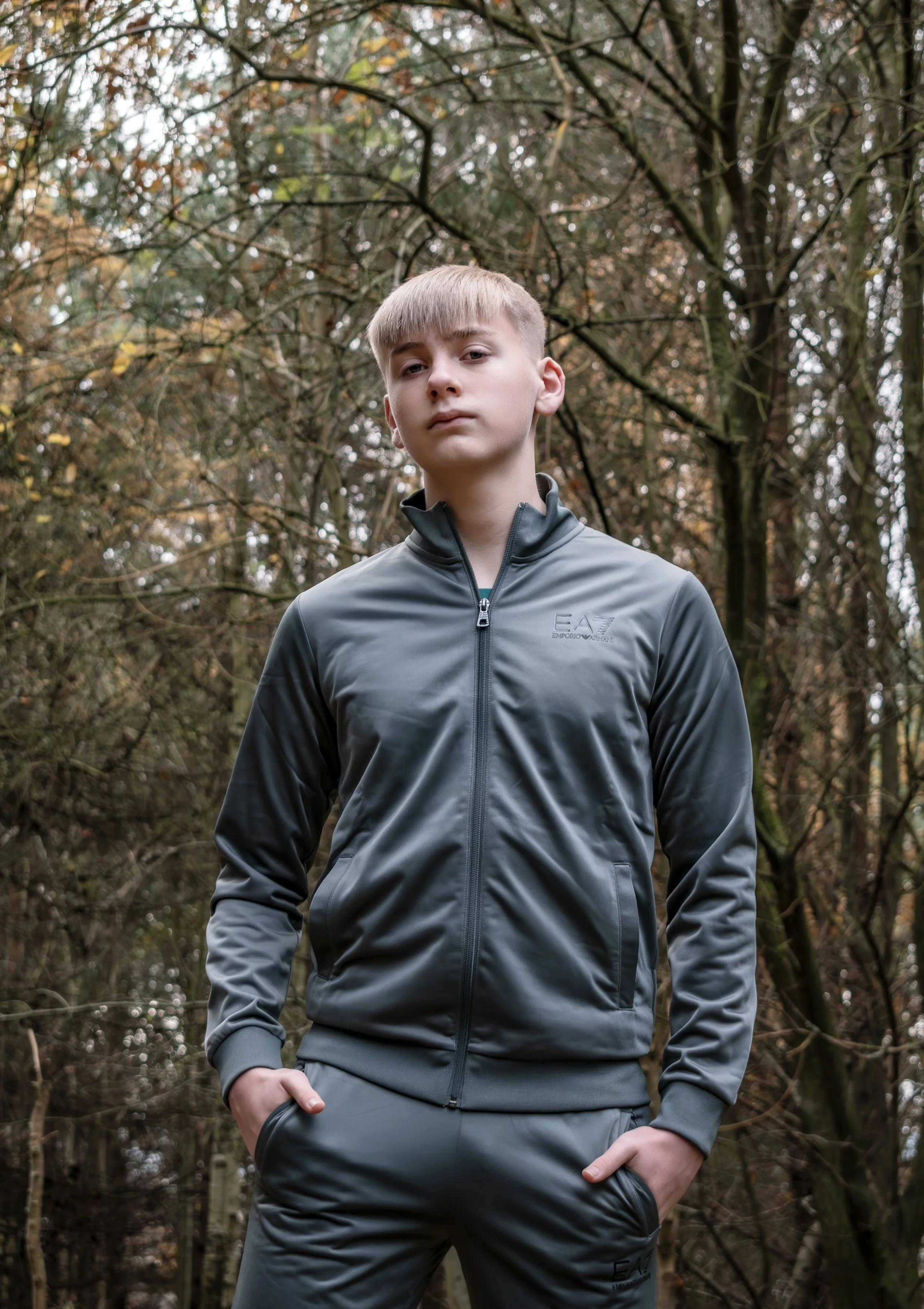 Young man standing outdoors in front of trees with autumn leaves, wearing a gray EA7 sports jacket and matching sweatpants, with hands in pockets and a confident expression.