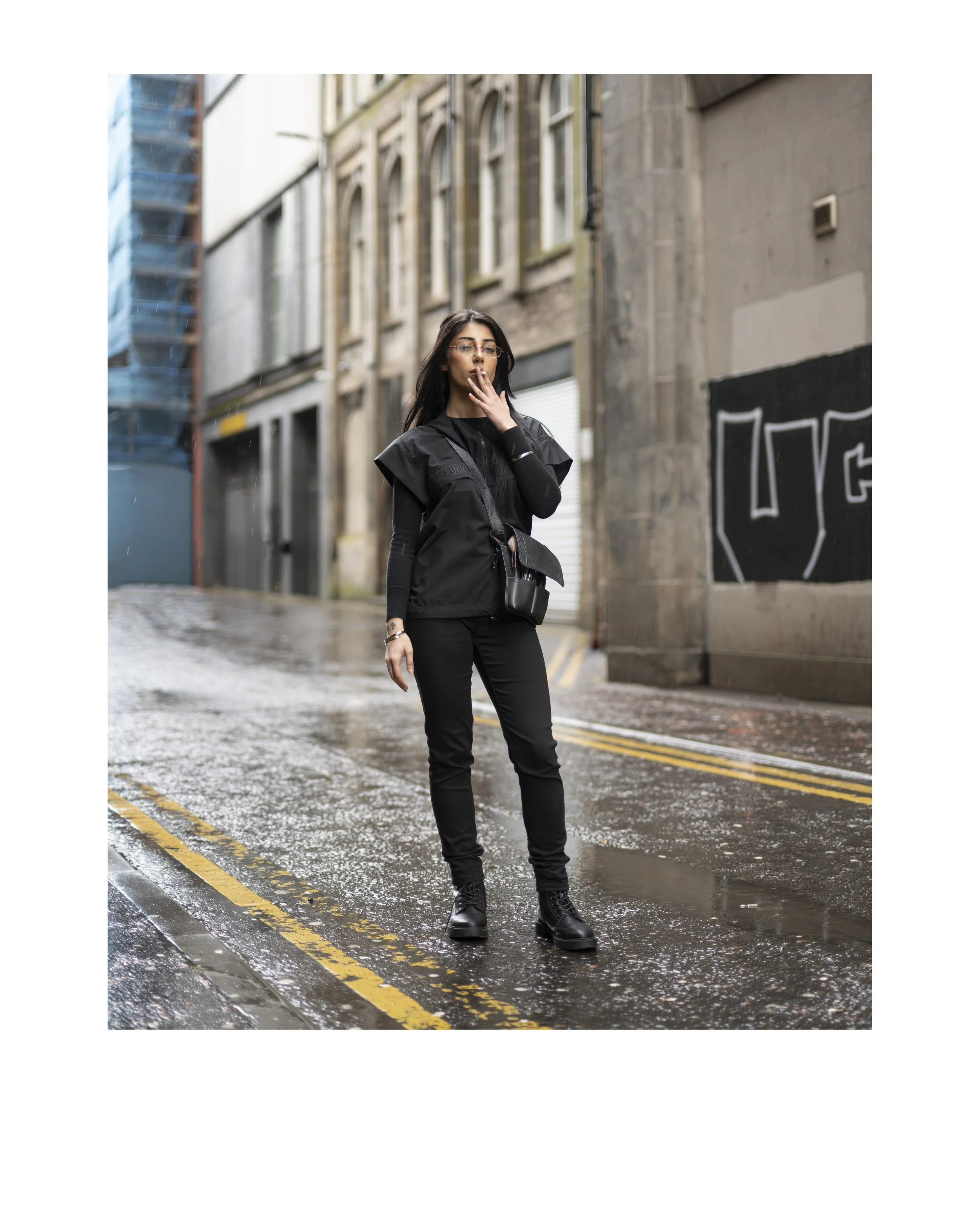 A woman in black clothing standing on a wet street with yellow road markings, overcast sky, city buildings in the background, and graffiti on the wall nearby.