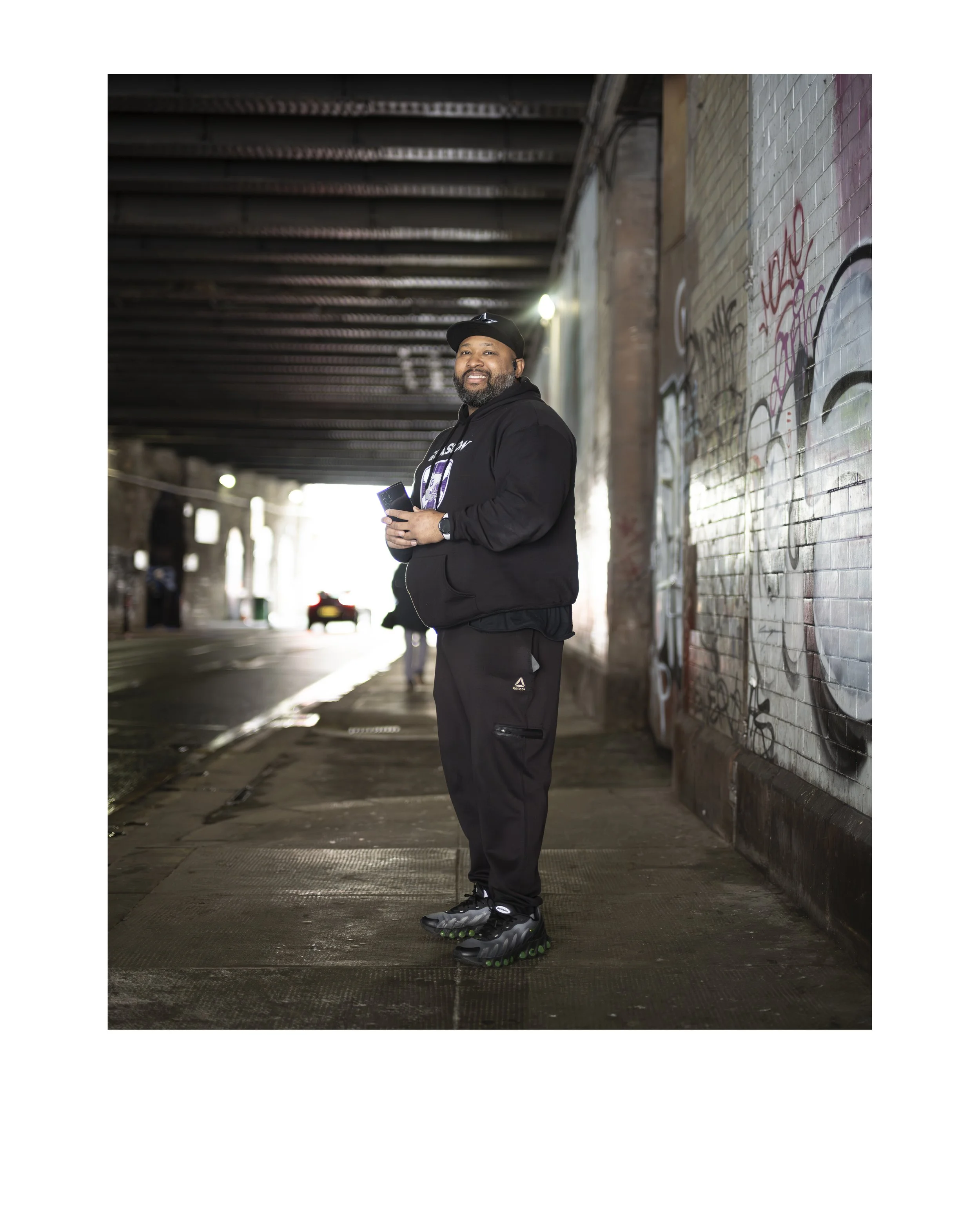 A man standing outdoors under a bridge, smiling and looking at the camera, holding a phone, wearing a black cap, hoodie, gray pants, and black sneakers with green soles, with graffiti on the brick wall to his right.