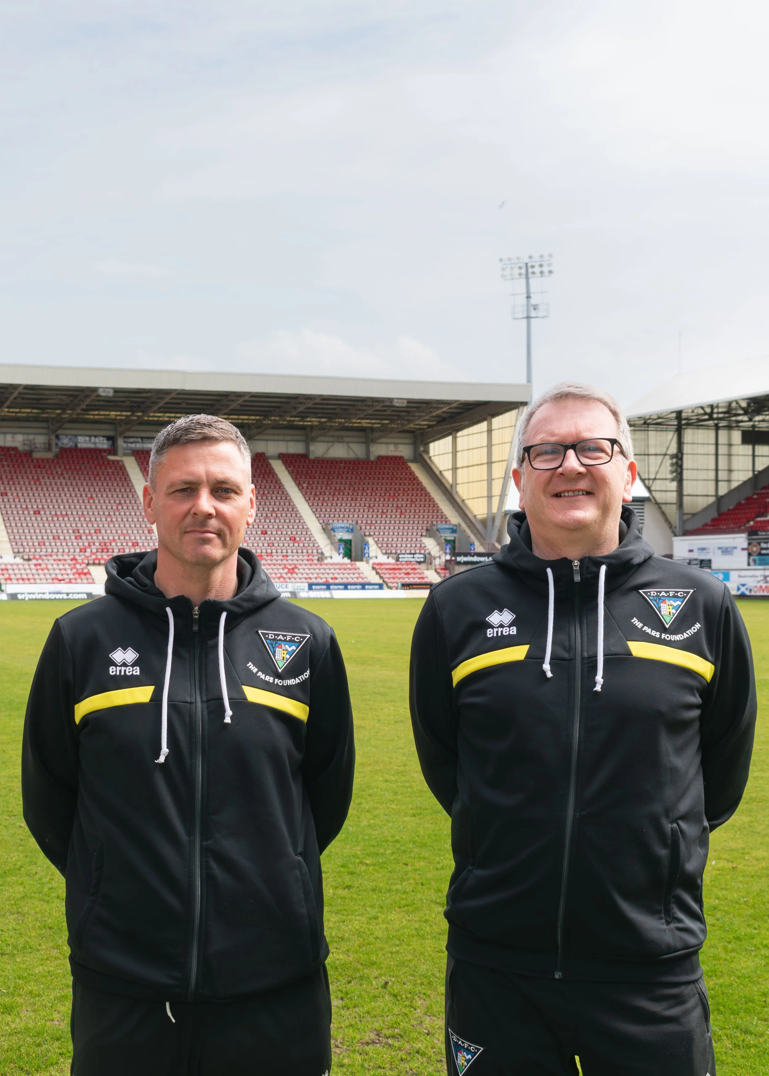Two men in black and yellow sports jackets standing on a football field in front of stadium seating, with one man wearing glasses and both smiling.