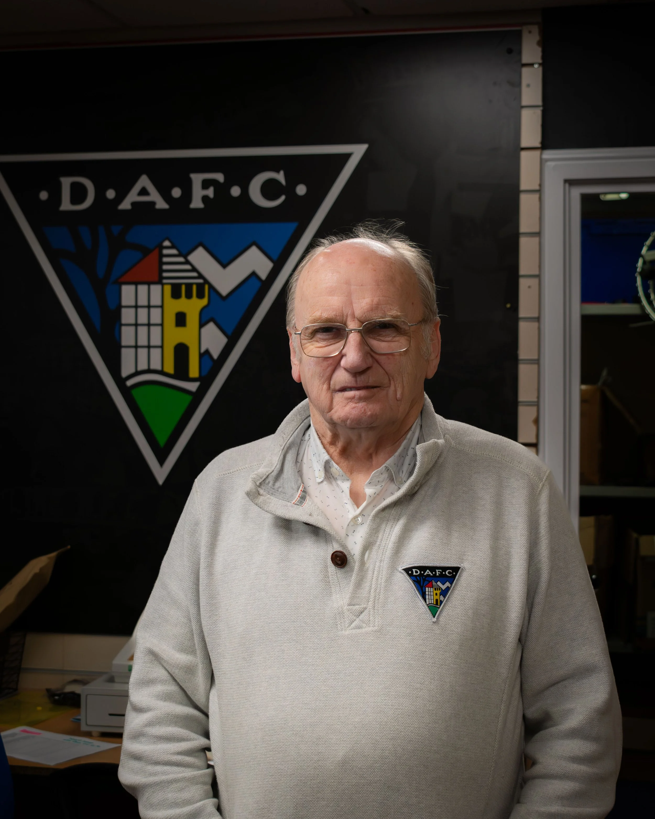 A senior man with glasses wearing a light-colored sweater with a DAFC emblem stands in front of a black wall with a DAFC logo. The background shows an indoor setting with a window and some office furniture.