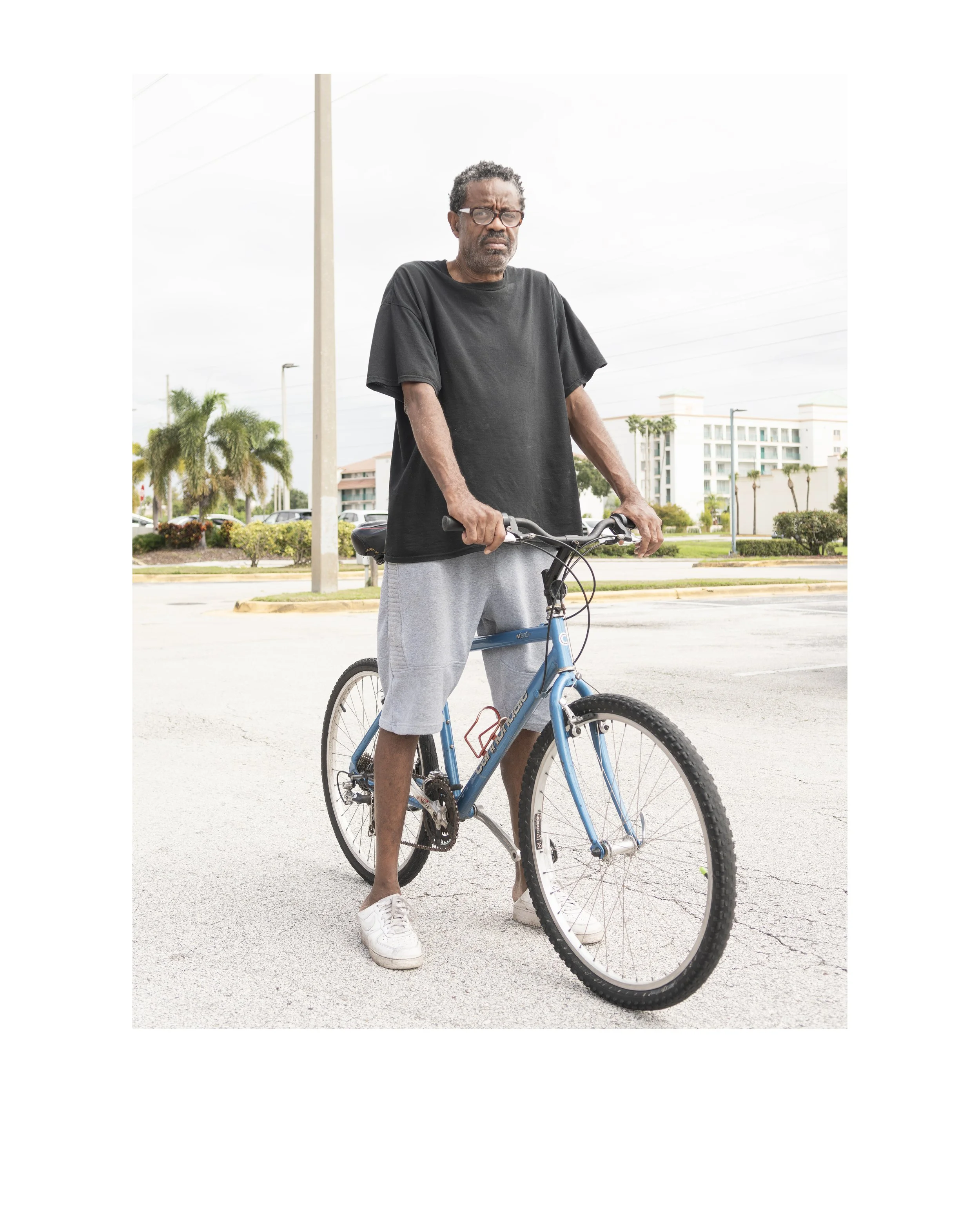 An elderly man with glasses and gray hair, wearing a black T-shirt and gray shorts, standing outdoors with his hand on a blue mountain bike, with palm trees and a multi-story building in the background.
