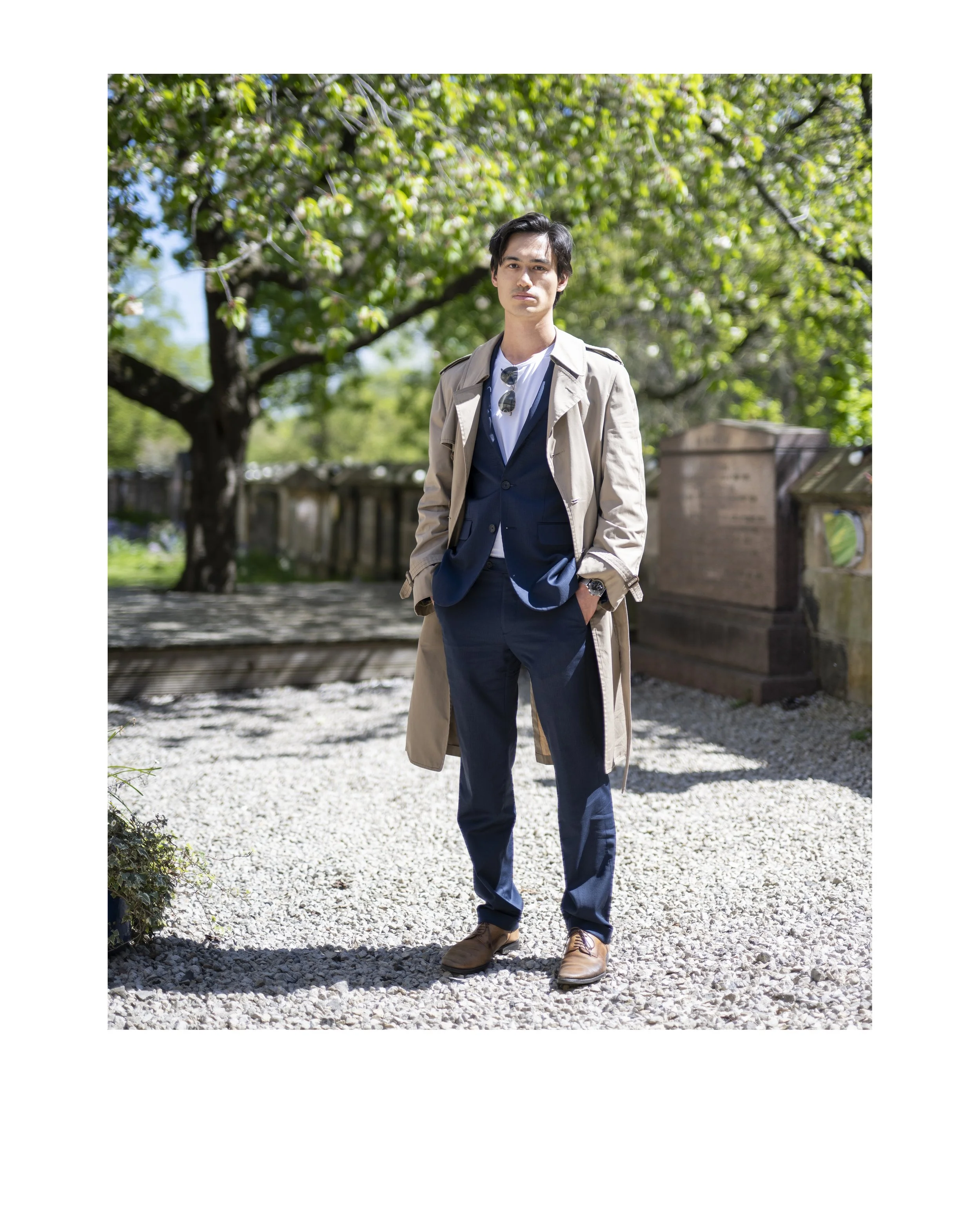 A young man standing outdoors on a sunny day with trees and a stone monument in the background, wearing a tan trench coat, dark suit, white shirt, sunglasses hanging from his shirt collar, and brown shoes.