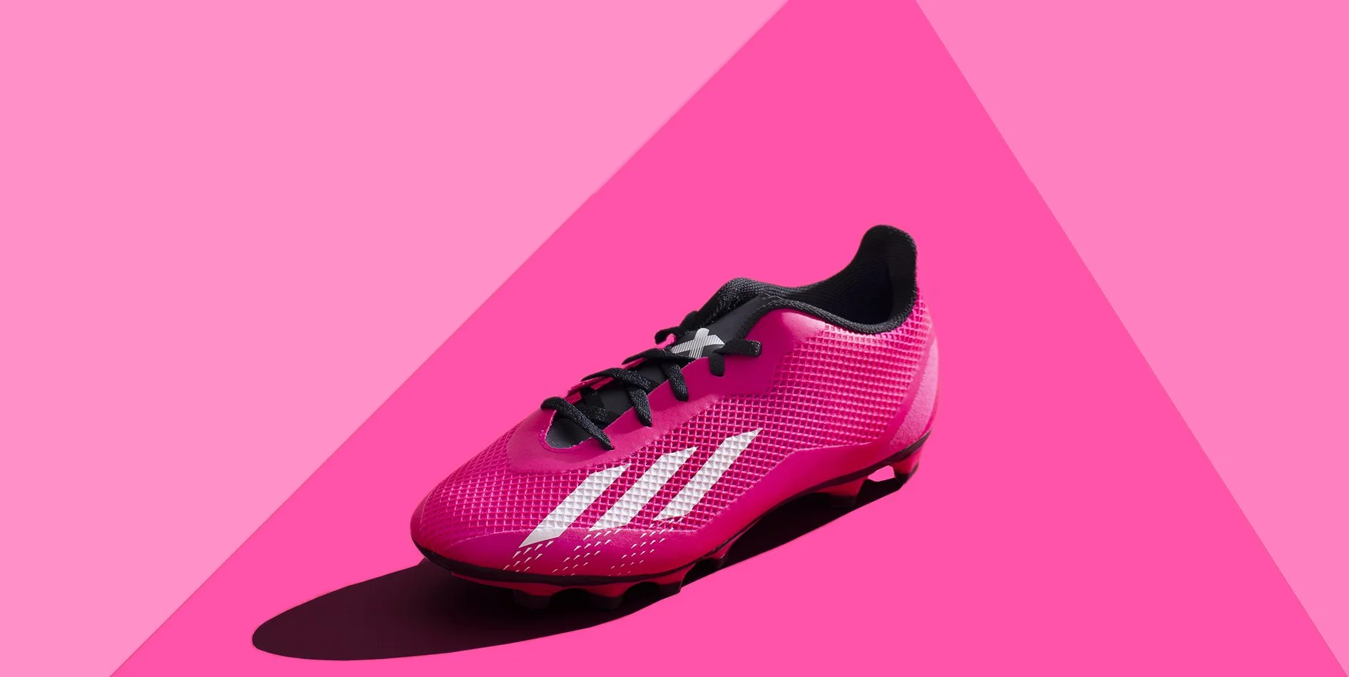 A pink and black Adidas soccer cleat with white stripes on a pink background with a shadow beneath.