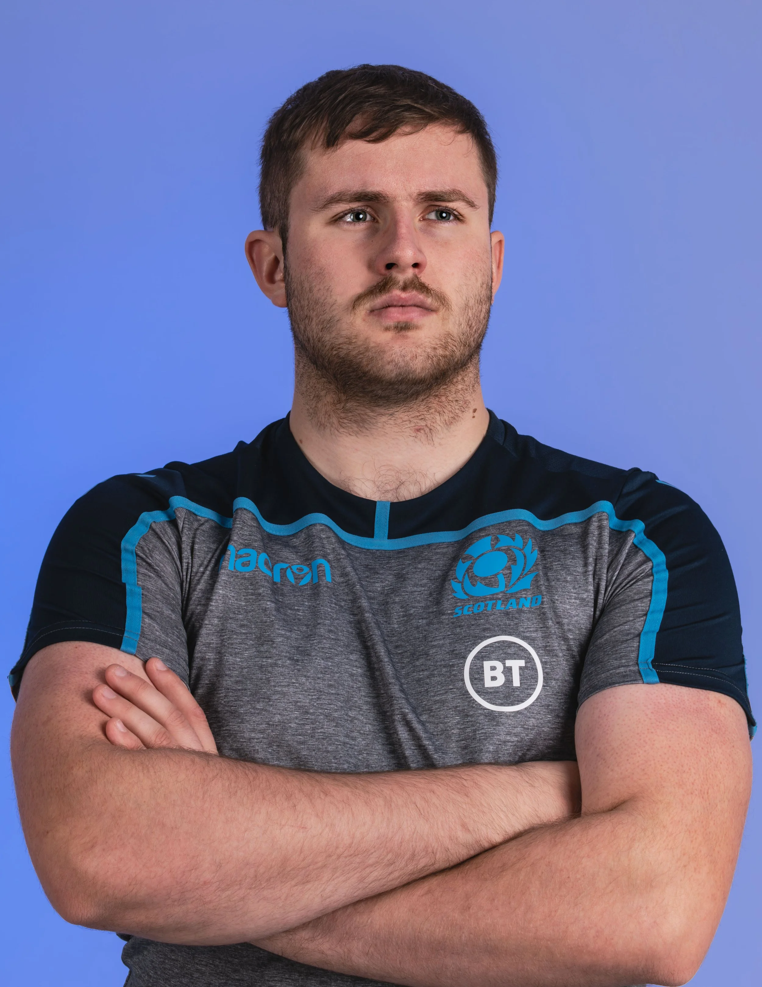 A male athlete with crossed arms, wearing a dark gray shirt with blue accents, featuring the Scotland rugby team logo and the BT sponsor logo, against a plain blue background.