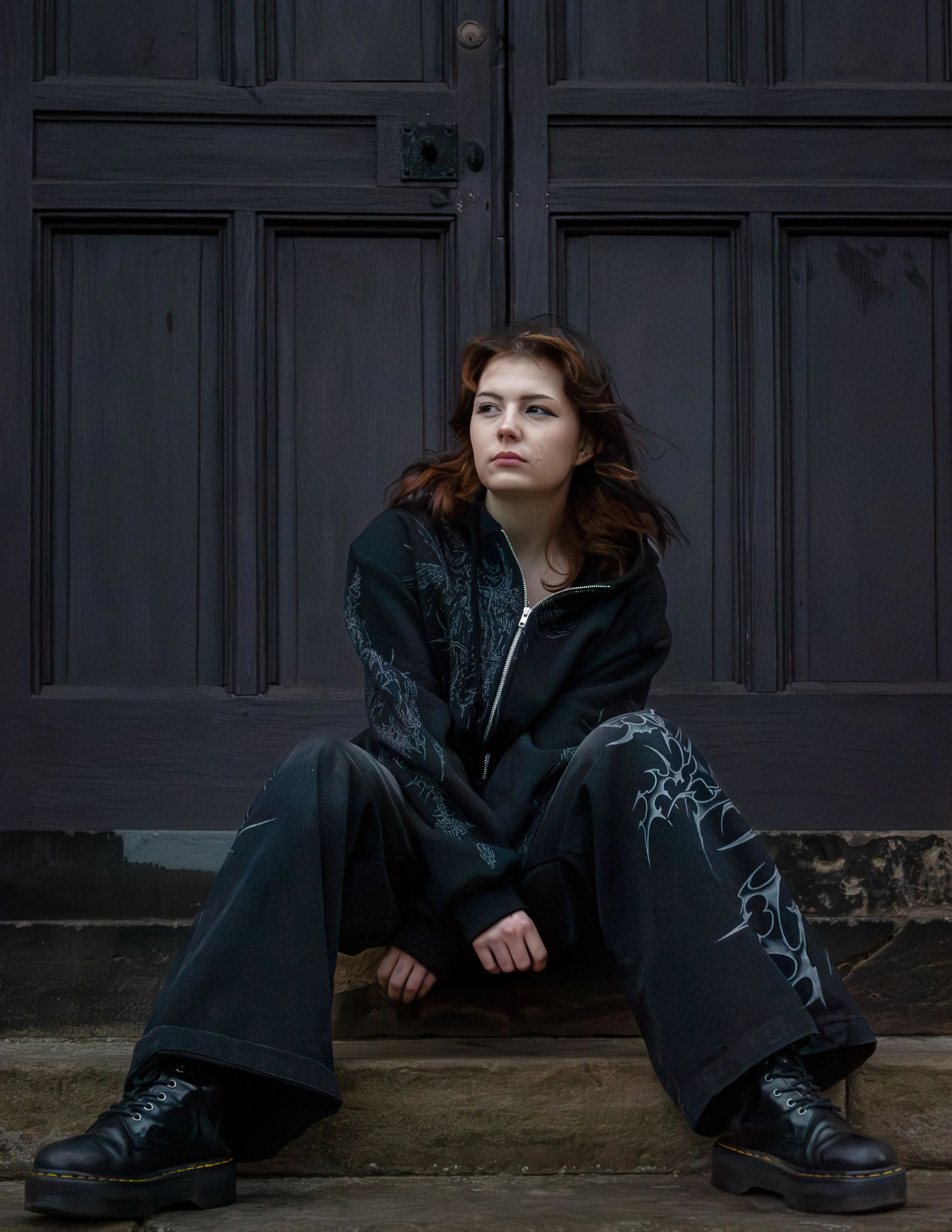 A young woman with wavy brown hair sitting in front of a large dark wooden door, wearing a black jacket with intricate designs, black pants, and black boots. She has a serious expression and is looking to her right.