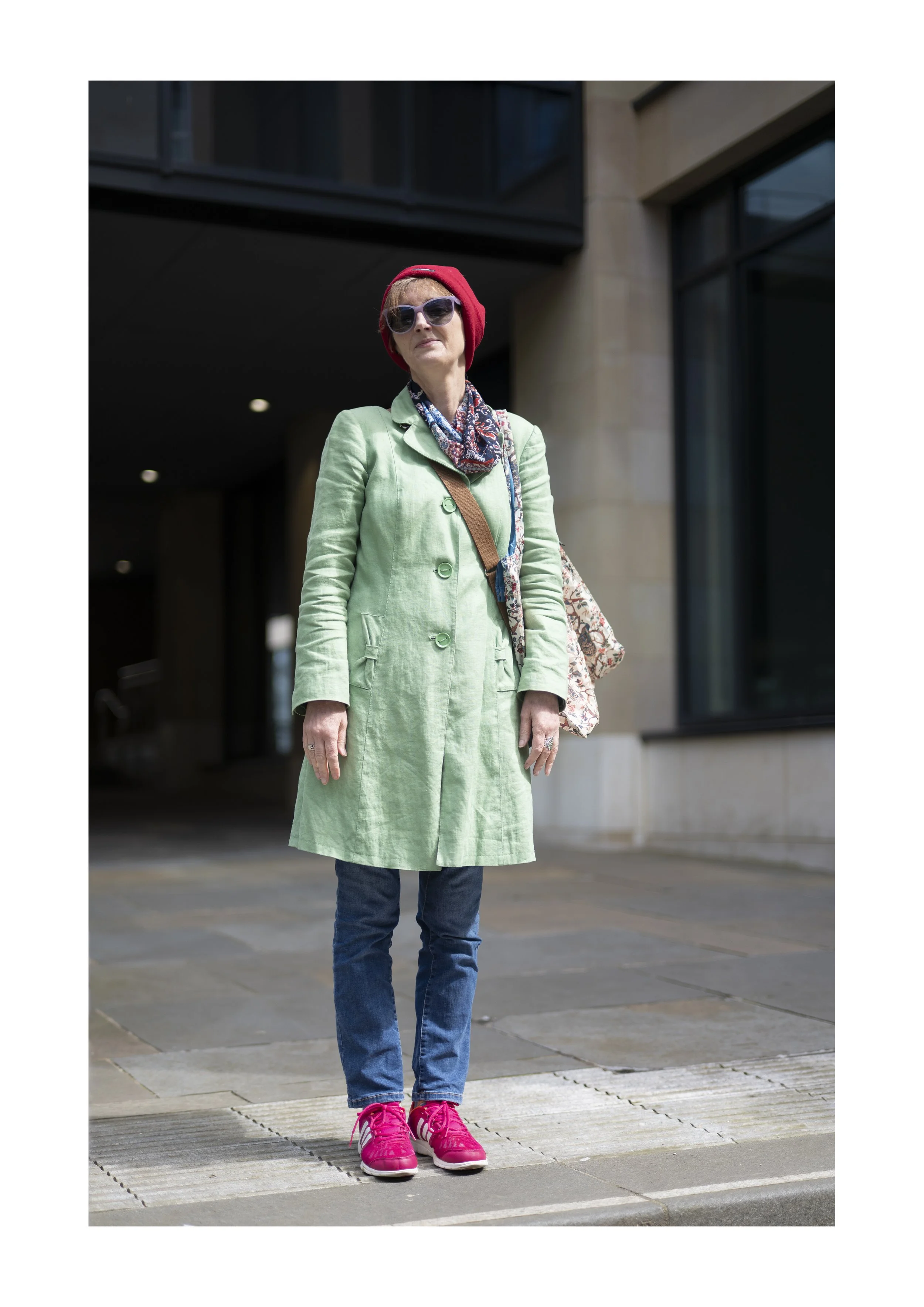 Close-up of a woman standing outdoors on a city sidewalk, wearing sunglasses, a red beanie, a green coat, jeans, and pink sneakers.