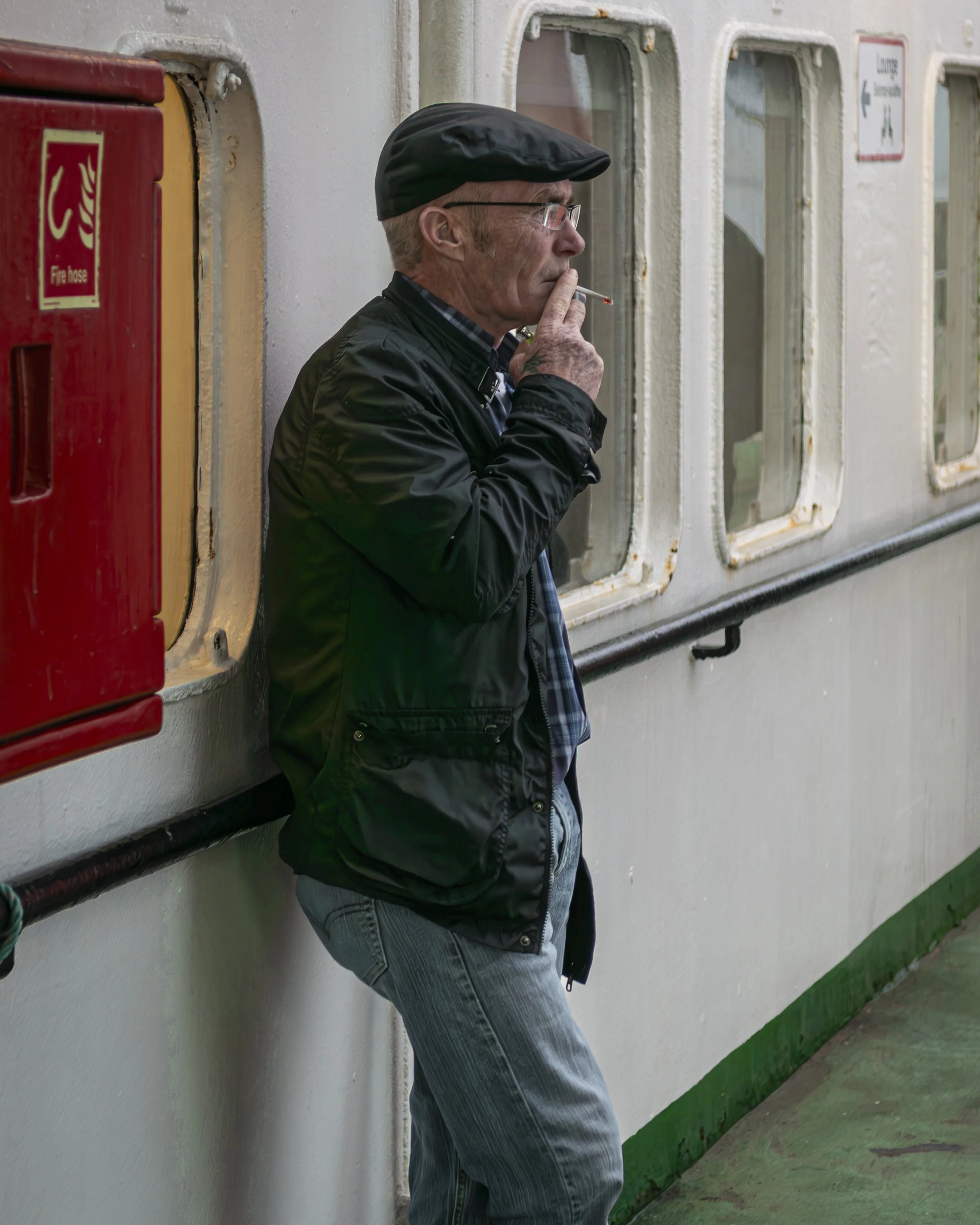 An elderly man with glasses and a black cap leaning against a wall, smoking a cigarette inside a building with worn paint and windows.