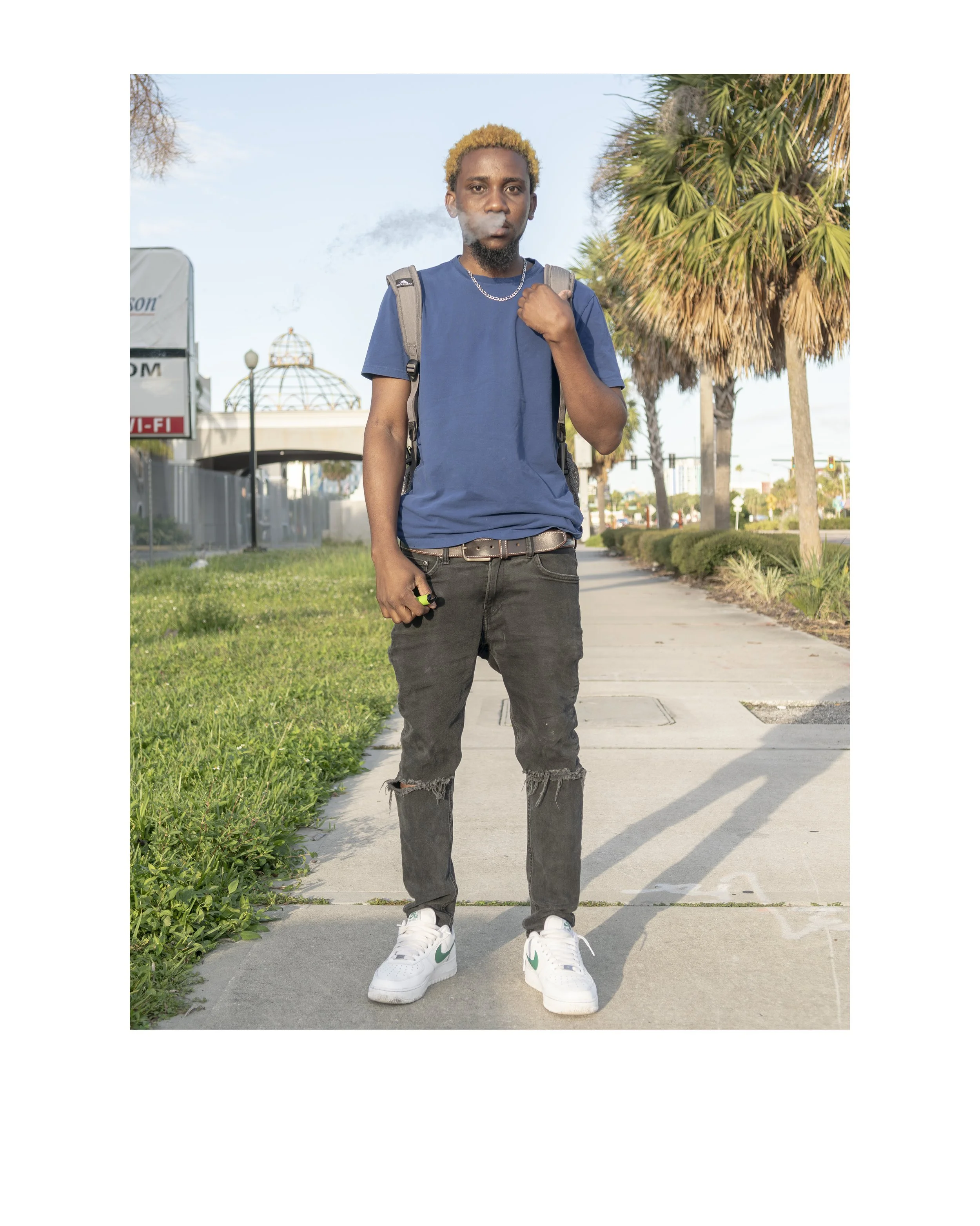 Young man with blonde hair, wearing a blue t-shirt, black ripped jeans, white sneakers, and carrying a backpack, standing outdoors on a sidewalk near palm trees with a clear blue sky in the background.