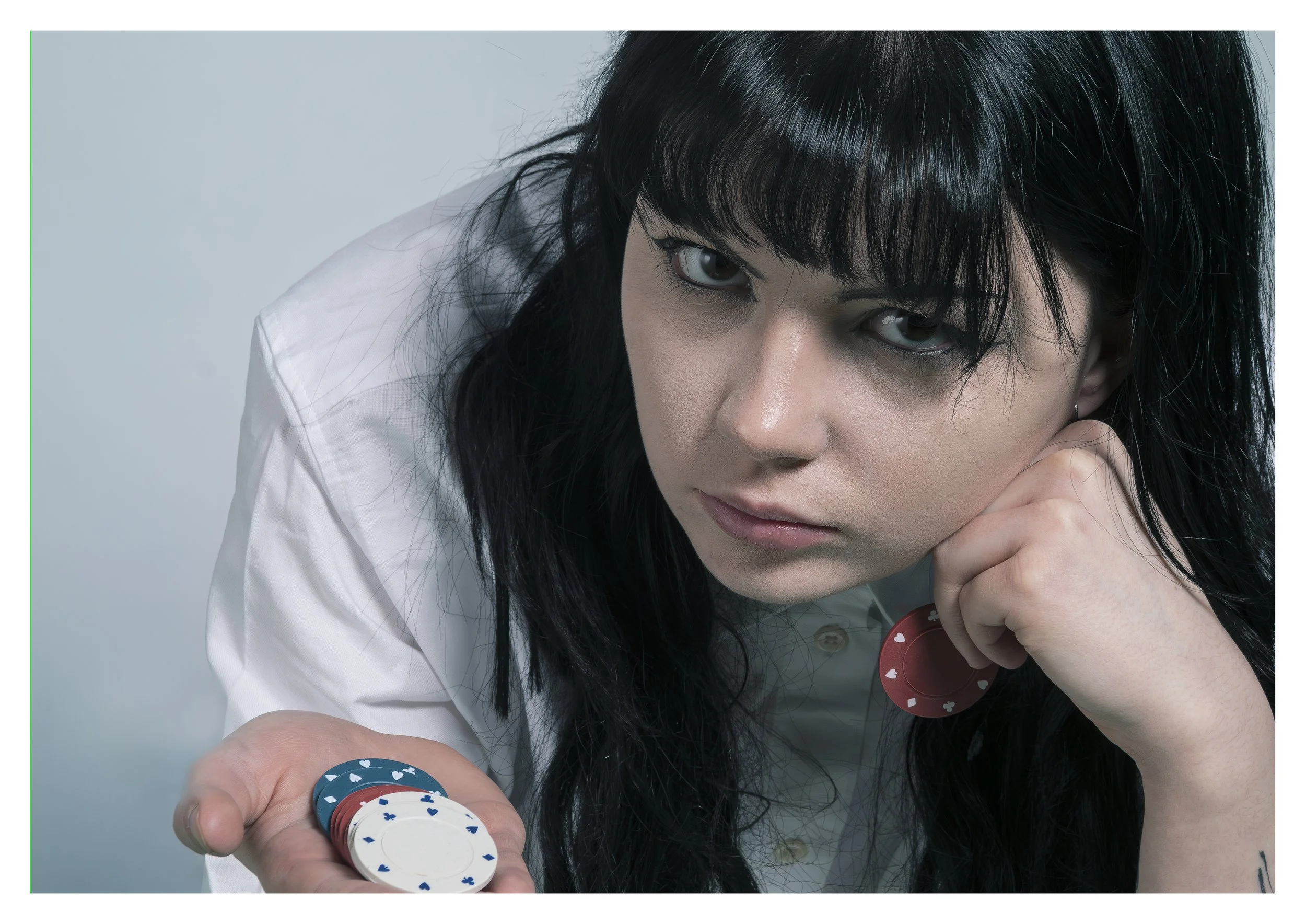 Young woman with black hair and bangs, wearing a white shirt, holding poker chips and looking up towards the camera against a light gray background.