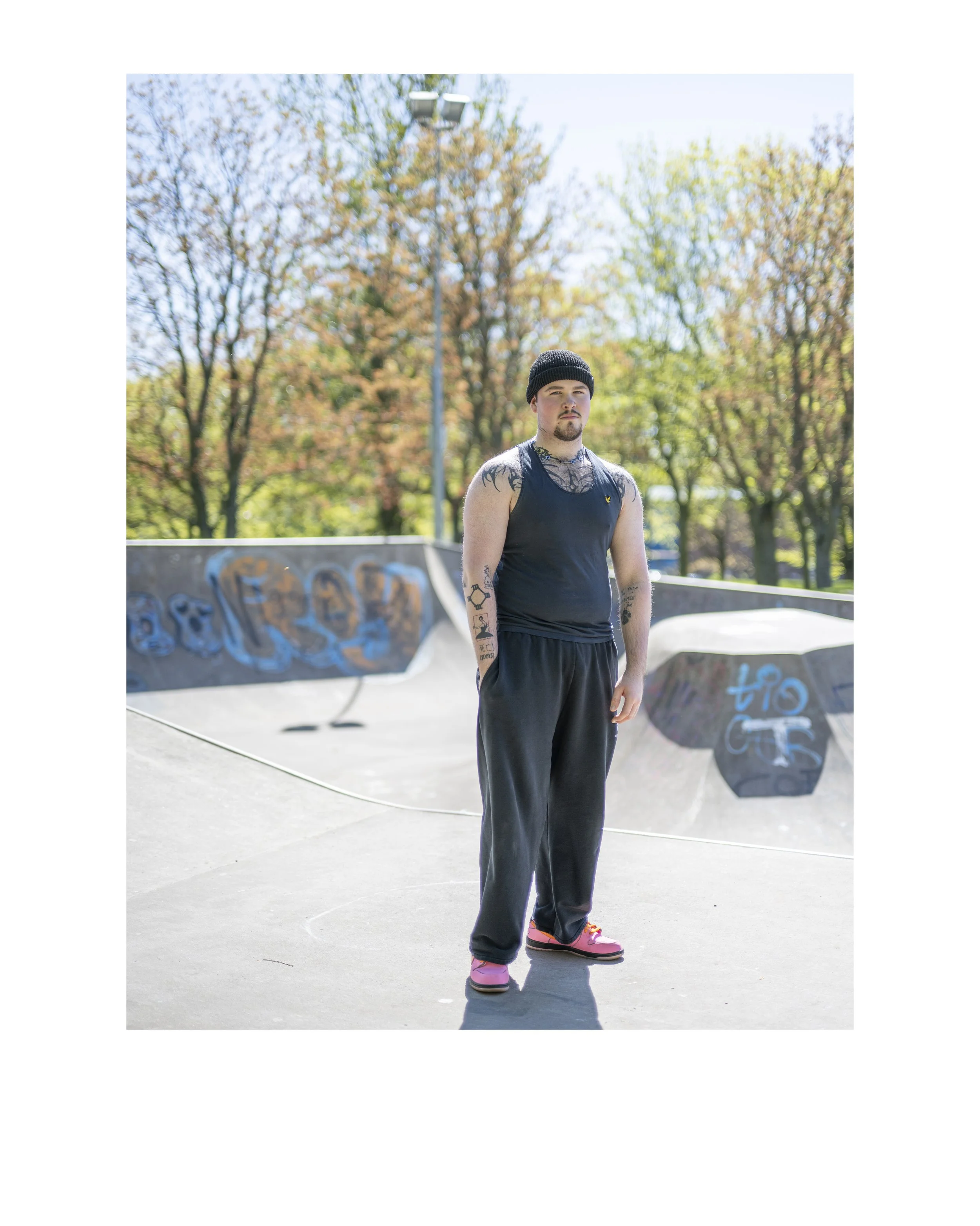 A man in a black sleeveless shirt and black sweatpants standing outdoors in a skate park with graffiti on the ramps, trees with autumn leaves, and a light pole in the background.