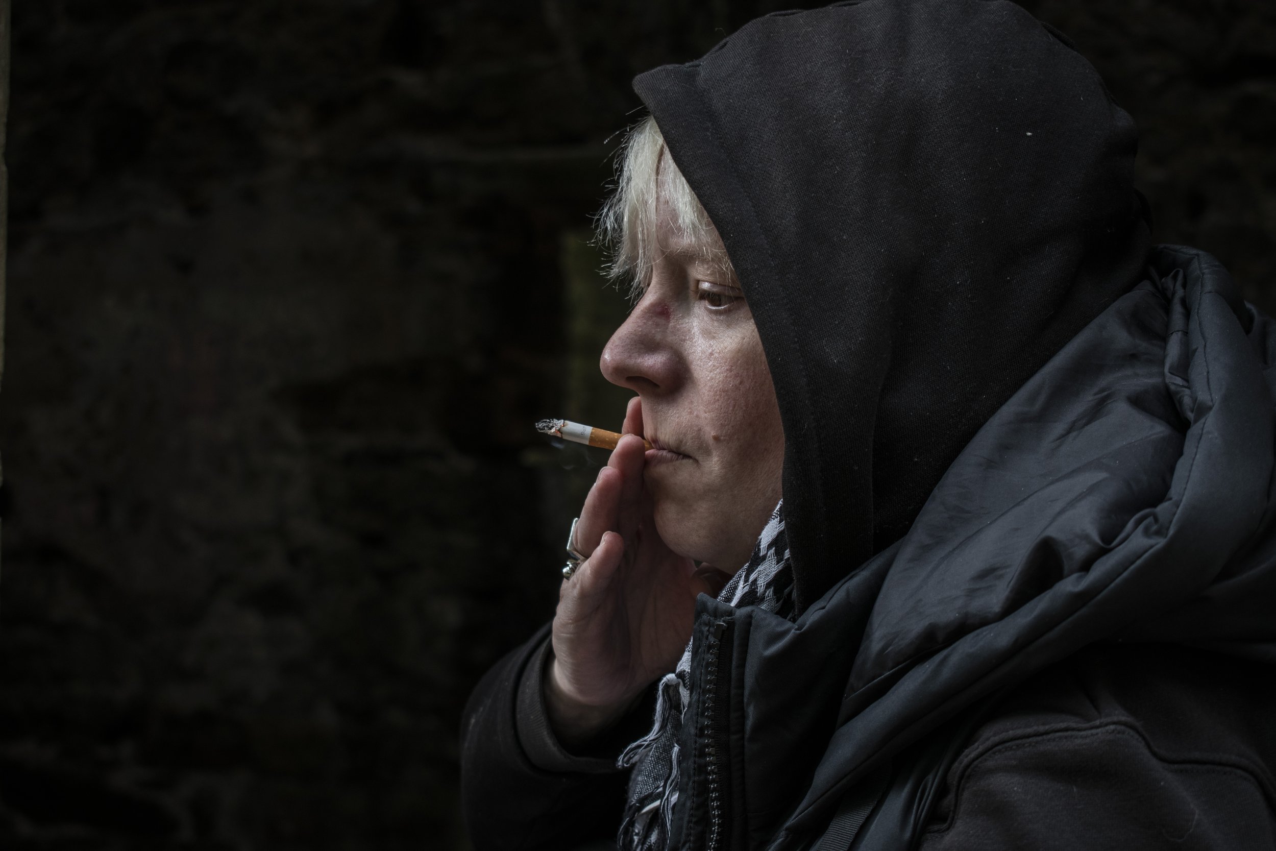 A person with light-colored hair wearing a dark jacket and hoodie, smoking a cigarette in a dark outdoor setting.