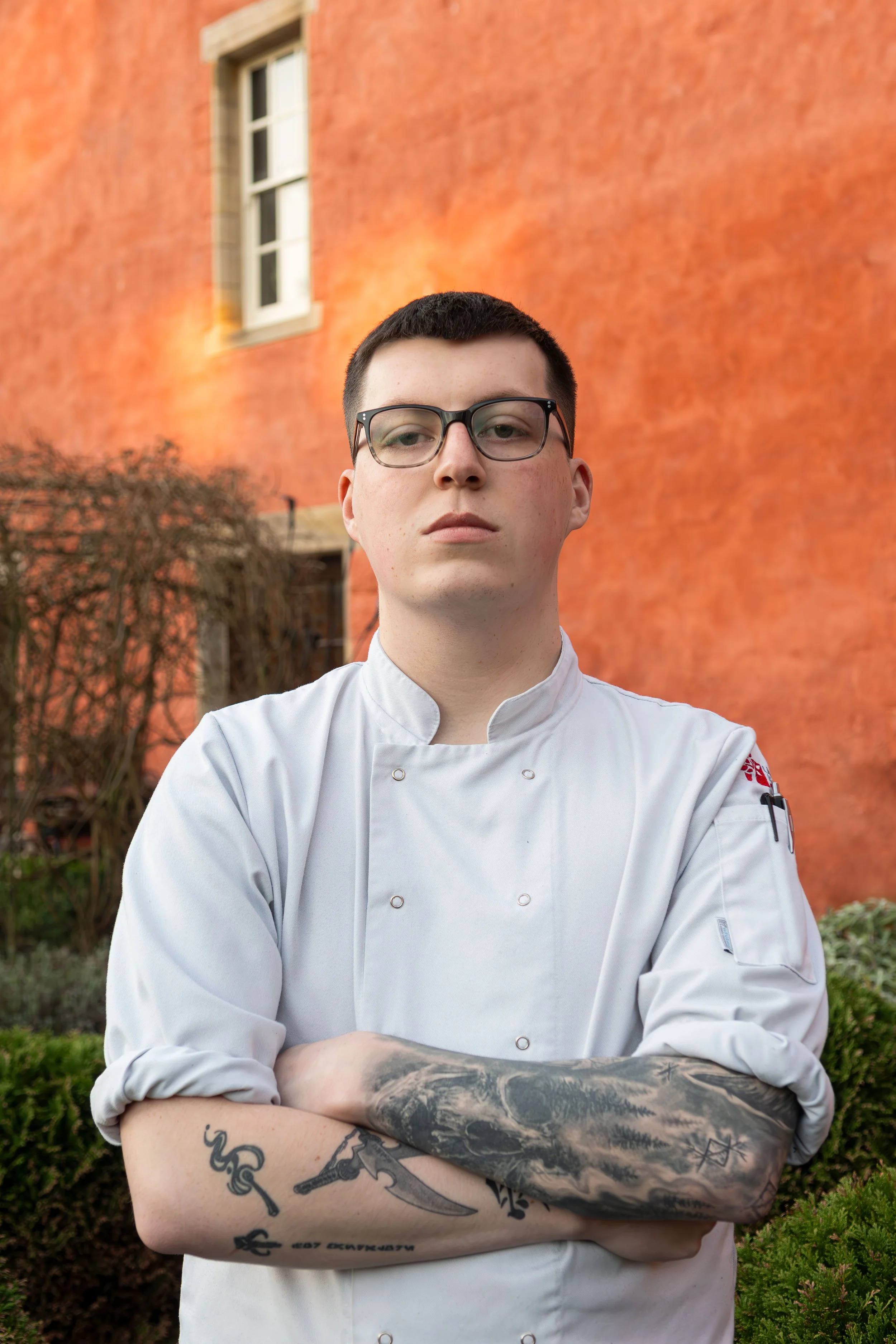 A young male chef with glasses, tattoos on his arms, wearing a white chef's coat, standing outdoors in front of an orange building with windows and some plants.
