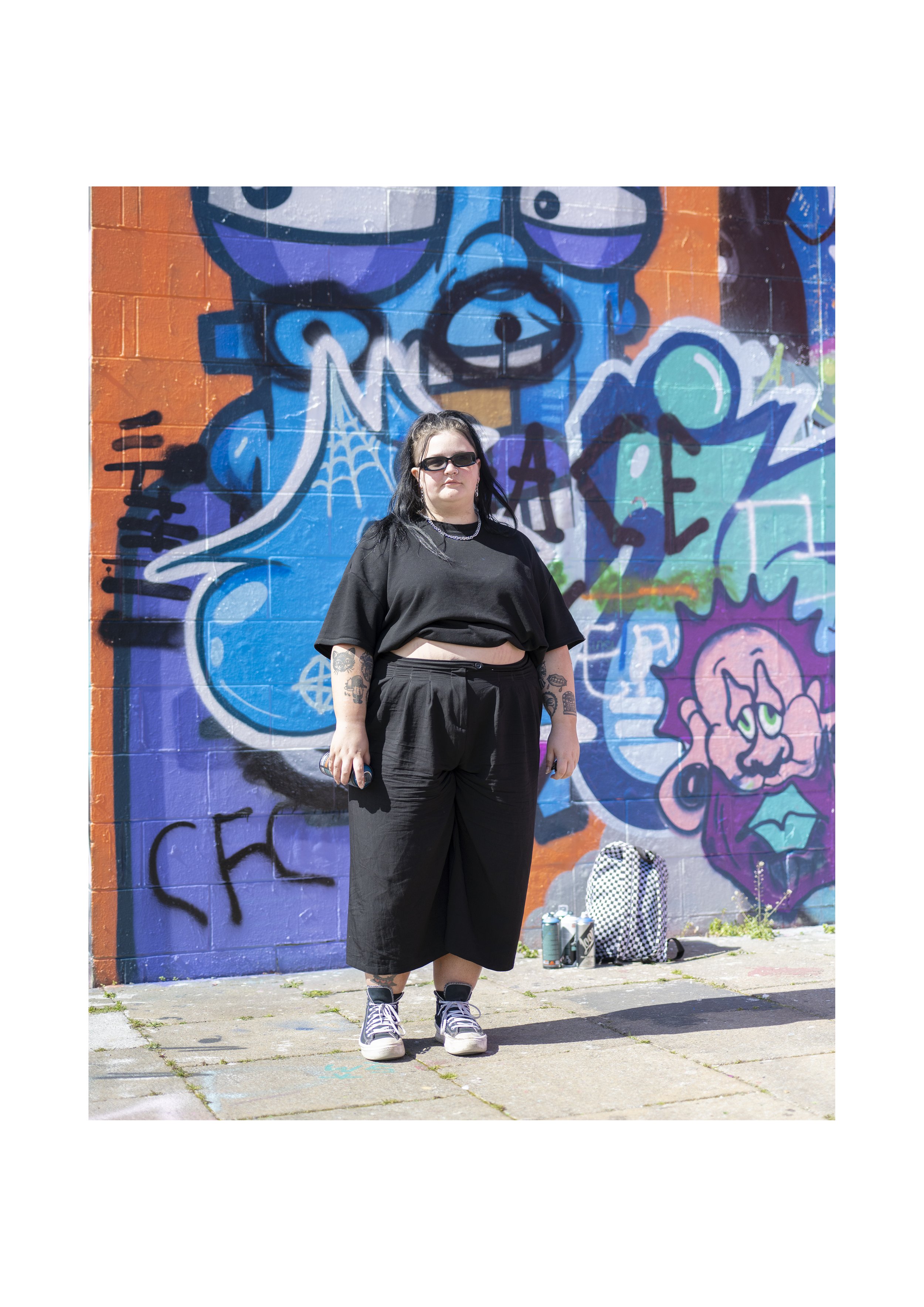 A woman with black hair, wearing sunglasses, a black crop top, and black pants, standing in front of colorful graffiti on a brick wall. There are spray paint cans and a checkered bag on the ground beside her.