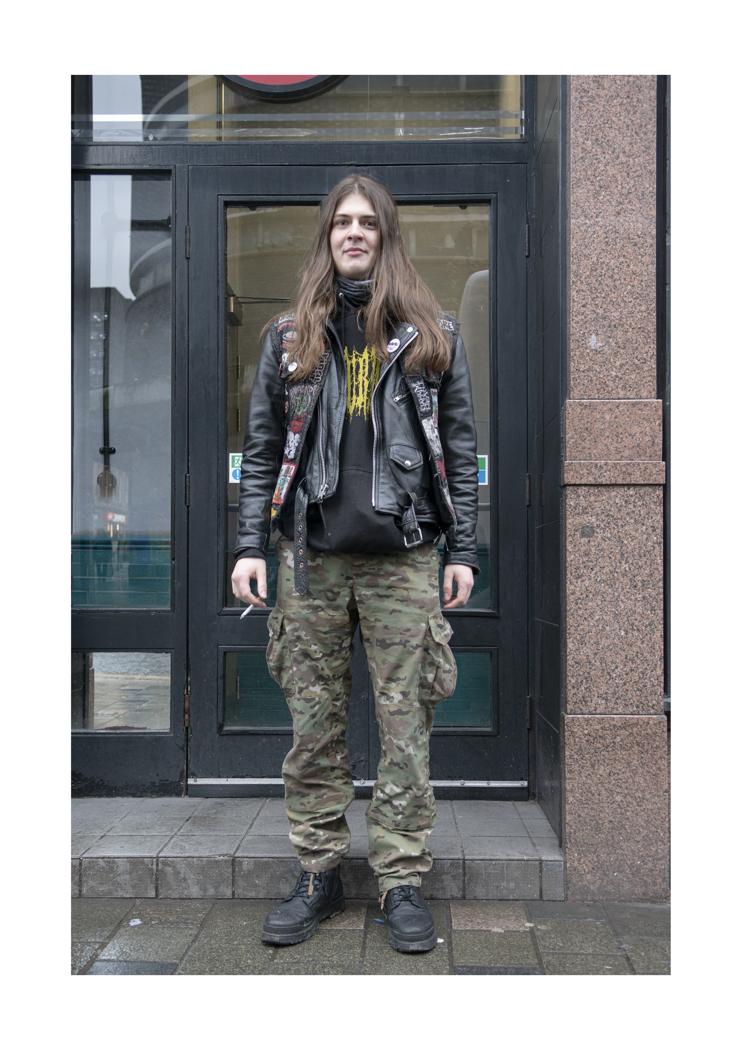 A young woman with long brown hair stands in front of a black door with glass panes. She is wearing a black leather jacket, a black T-shirt with yellow text, camouflage pants, and black combat boots.