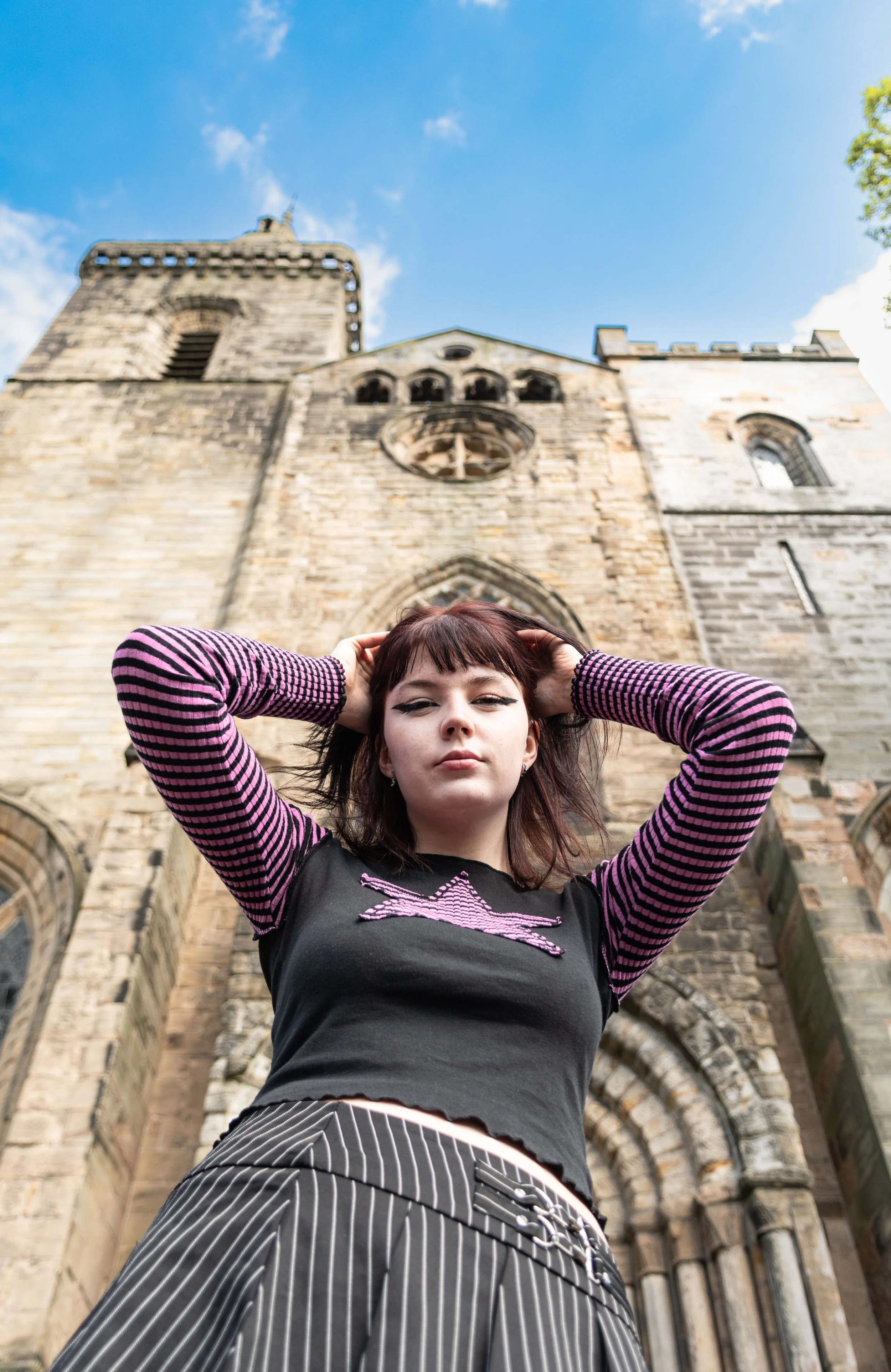A young woman with dark hair and bangs posing outdoors in front of a historic stone church, looking down at the camera with her hands on her head. She wears a black shirt with pink detailing and striped pants, against a bright blue sky.