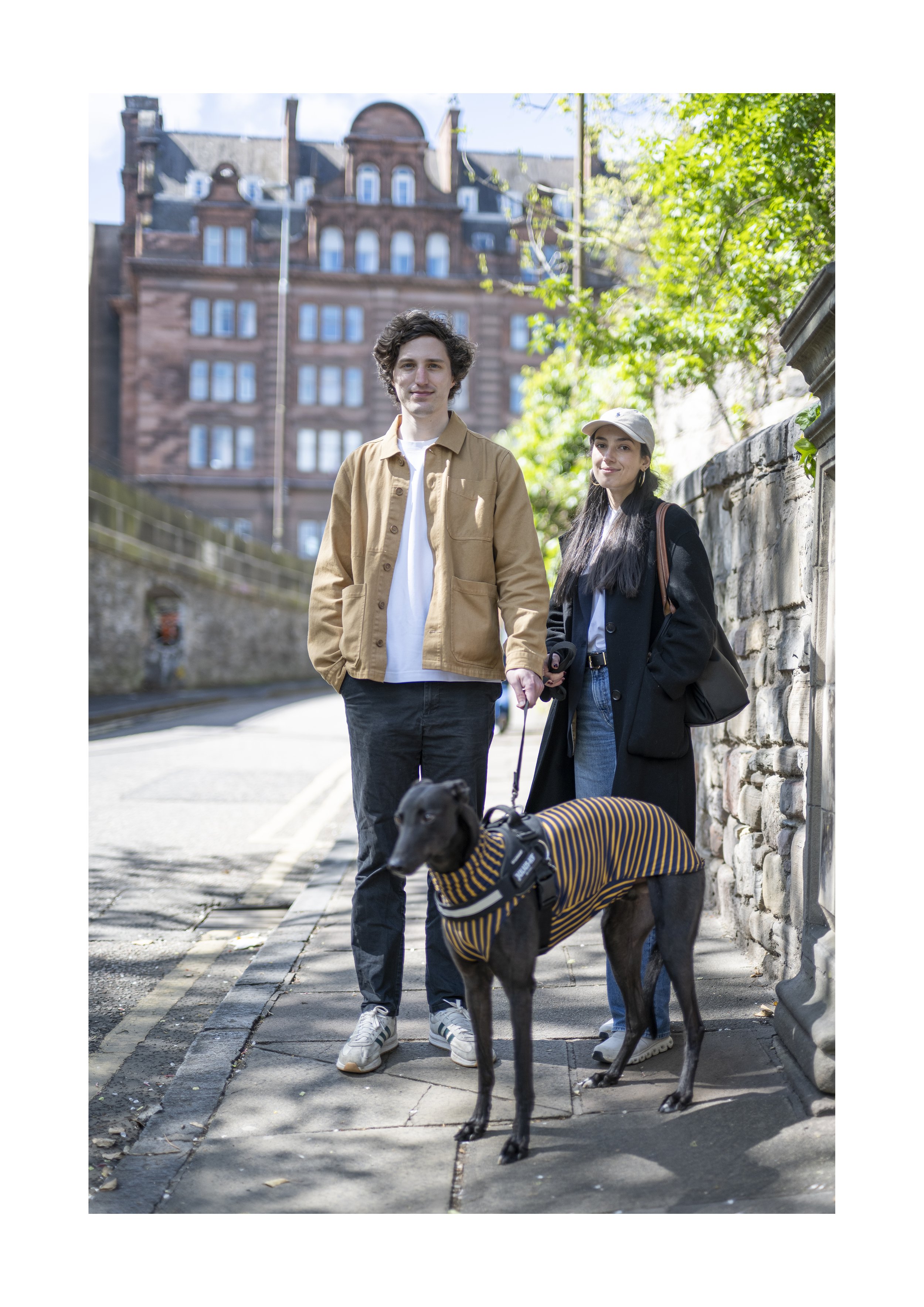 A young man and woman are walking a black dog on a city sidewalk. The dog is wearing a striped sweater, and the woman is dressed in a dark coat and white cap. The man has curly hair and is dressed in a tan jacket, while the woman has long dark hair a
