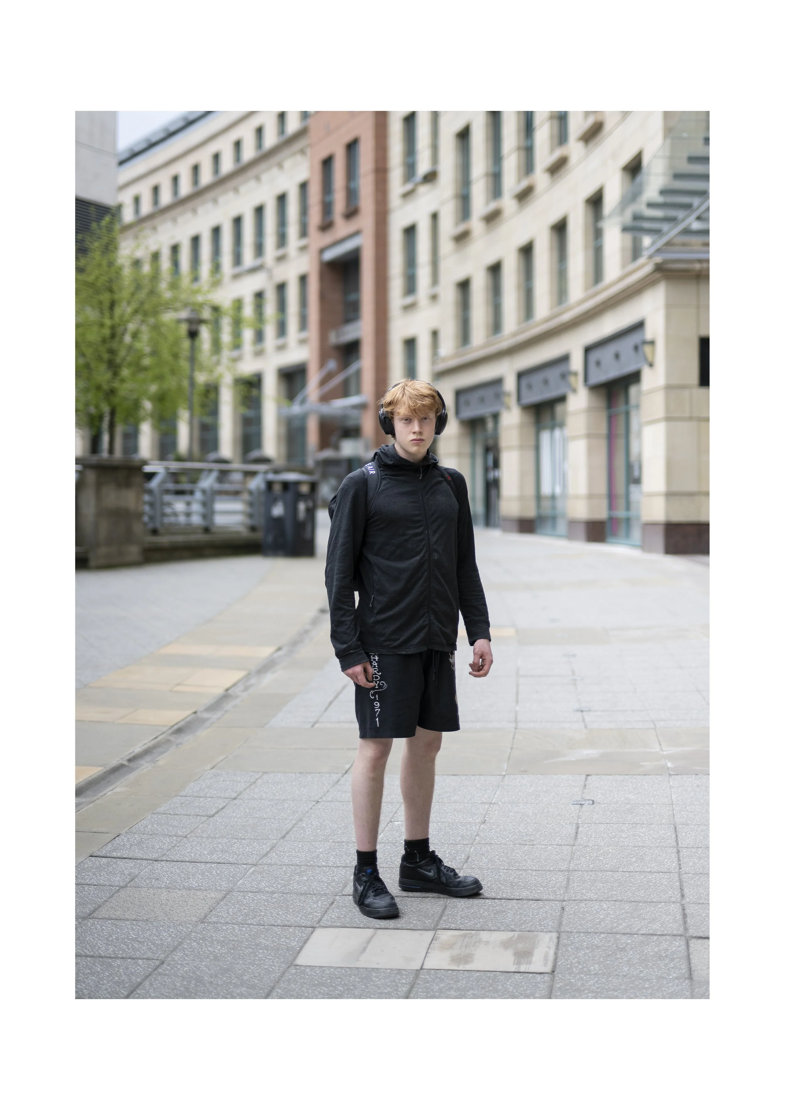A young man with blond hair wearing a black hoodie and shorts, black sneakers, black headphones, and a backpack standing on a city sidewalk with buildings in the background.