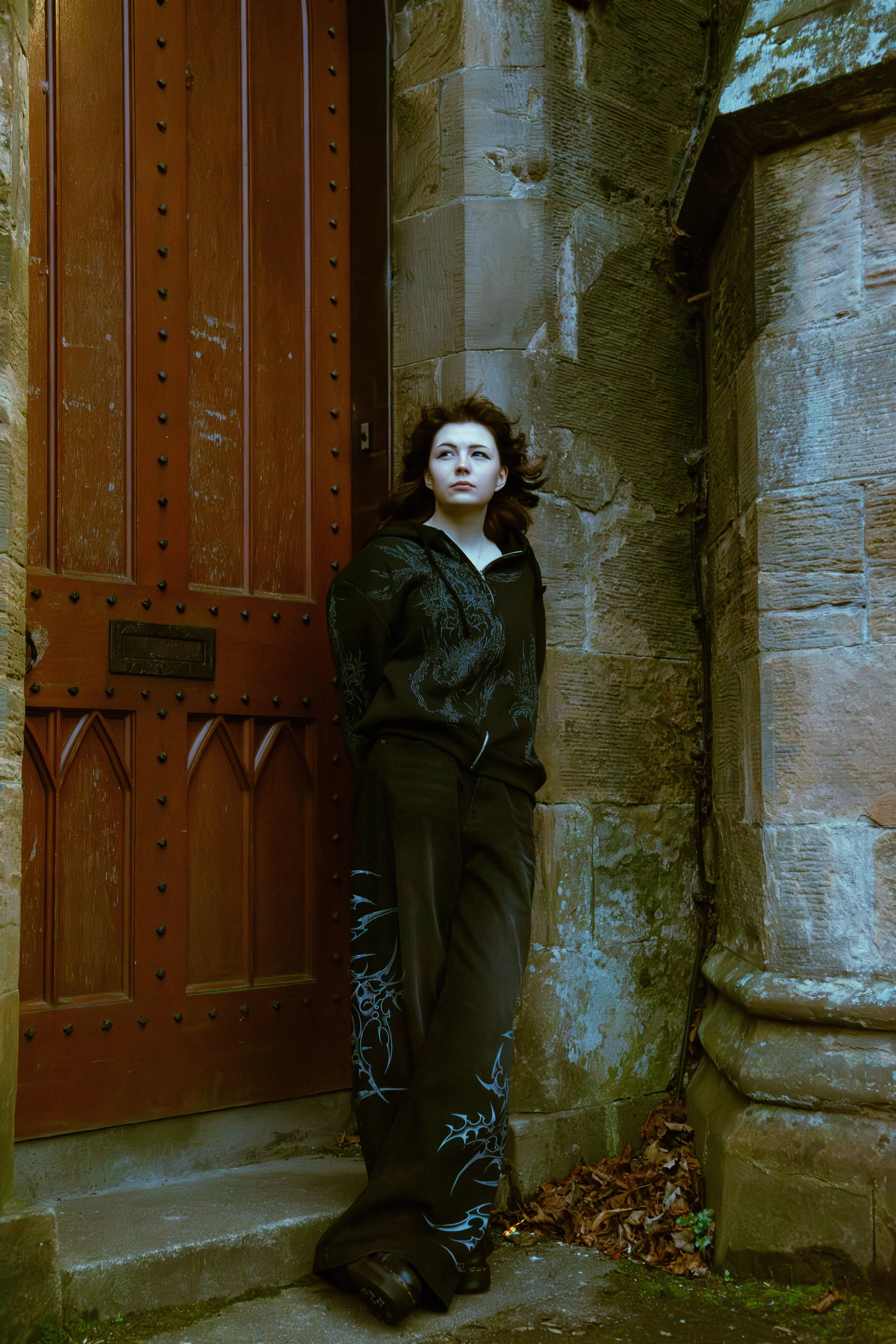 A young woman with dark hair standing outside near a wooden door and stone wall, wearing black clothing with blue and white designs.