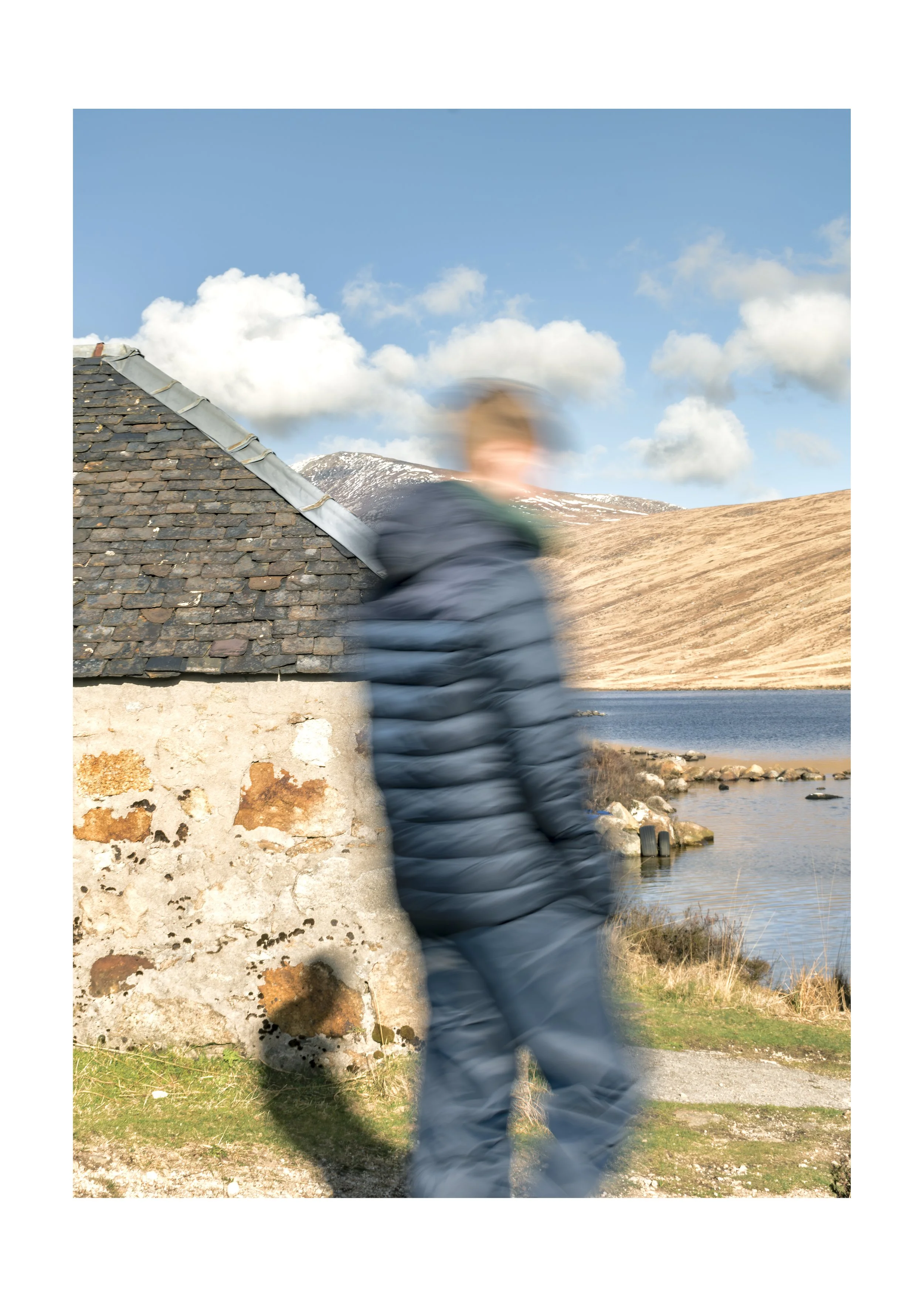 Blurry person in a puffer jacket walking near a stone building with a sloped roof, a lake, and mountains with snow in the background under a partly cloudy sky.