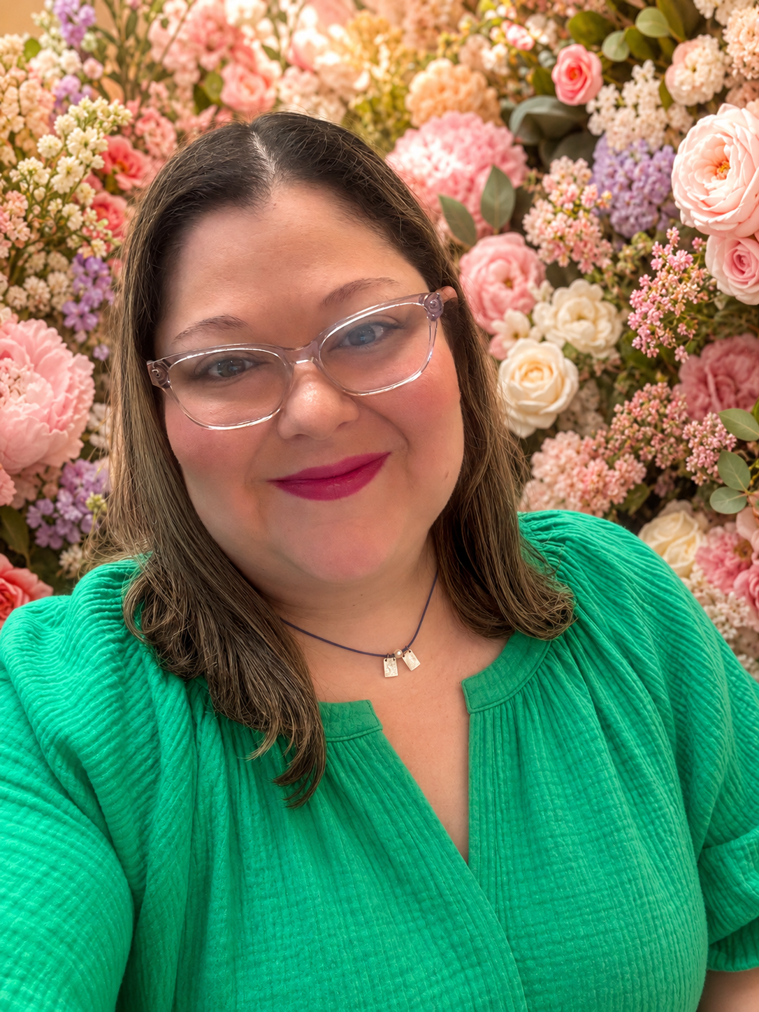 A smiling woman with shoulder-length brown hair, wearing clear glasses and a green top, standing in front of a wall of pink, white, and purple flowers.