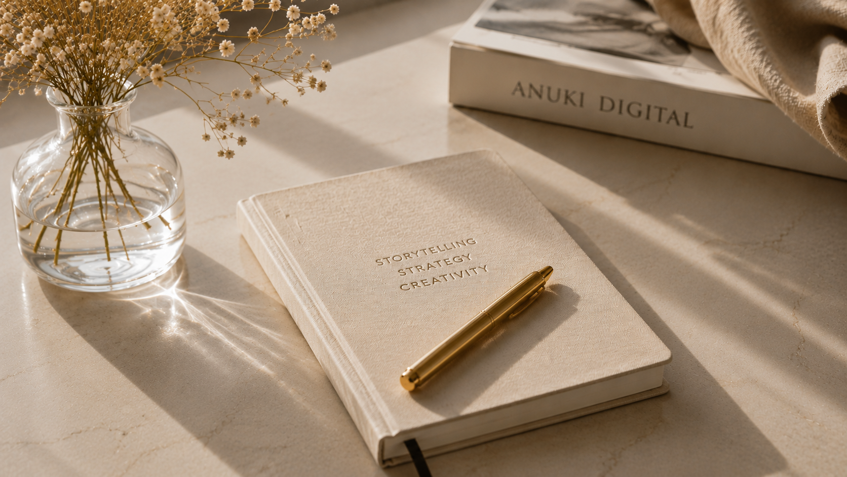 A beige notebook with the words 'Storytelling Strategy Creativity' embossed on the cover, a gold pen resting on top, a glass vase with dried white flowers to the left, a book titled 'Anuki Digital' in the background, and some fabric and papers nearby on a light-colored surface with soft sunlight.