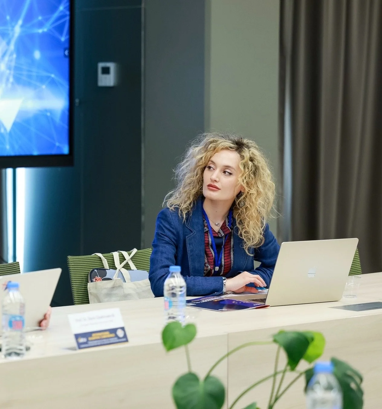 A young woman with curly blonde hair sits at a conference table with a laptop in front of her, wearing a denim jacket and a lanyard, in a professional setting.