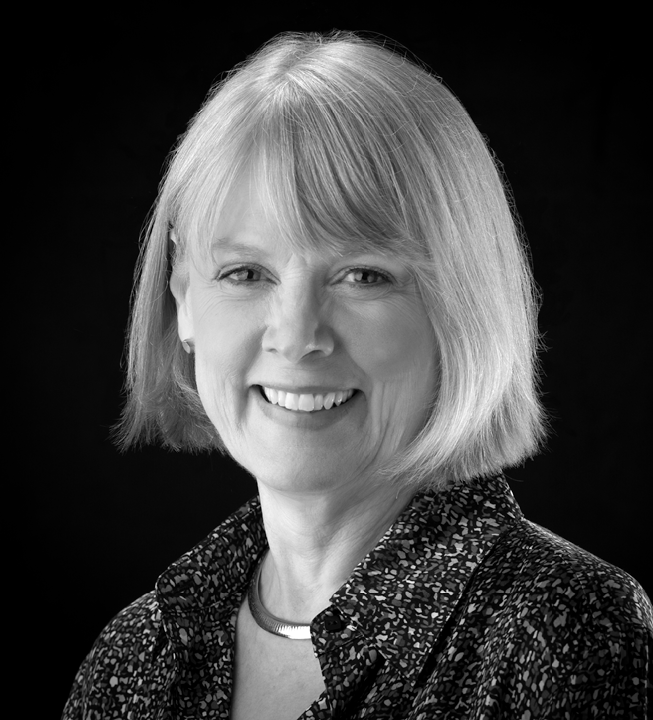 Black and white portrait of a smiling woman with shoulder-length hair, wearing a patterned blouse and a necklace, against a dark background.