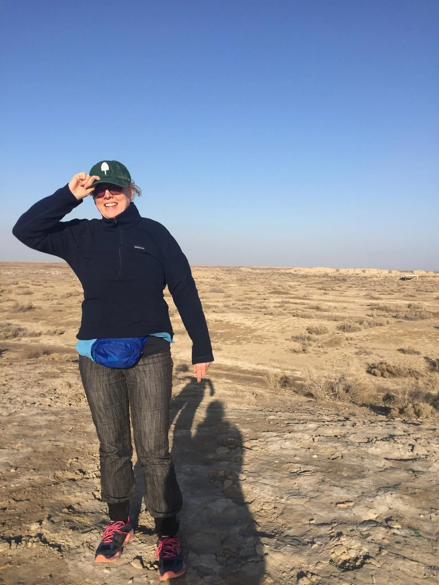 A woman smiling and wearing sunglasses, a green cap, a black jacket, gray pants, and pink and black running shoes, standing in a dry, open landscape under a clear blue sky.