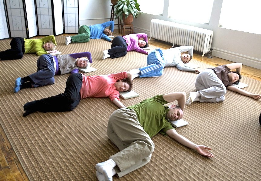 Multiple people practicing yoga or stretching on the floor with pillows under their heads in a bright, room with large windows and a plant.