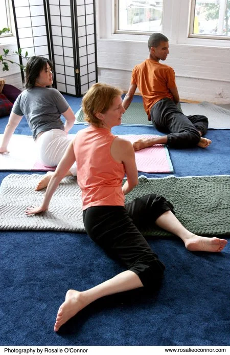 Three people practicing yoga or stretching on mats in a bright indoor space with large windows and white walls.