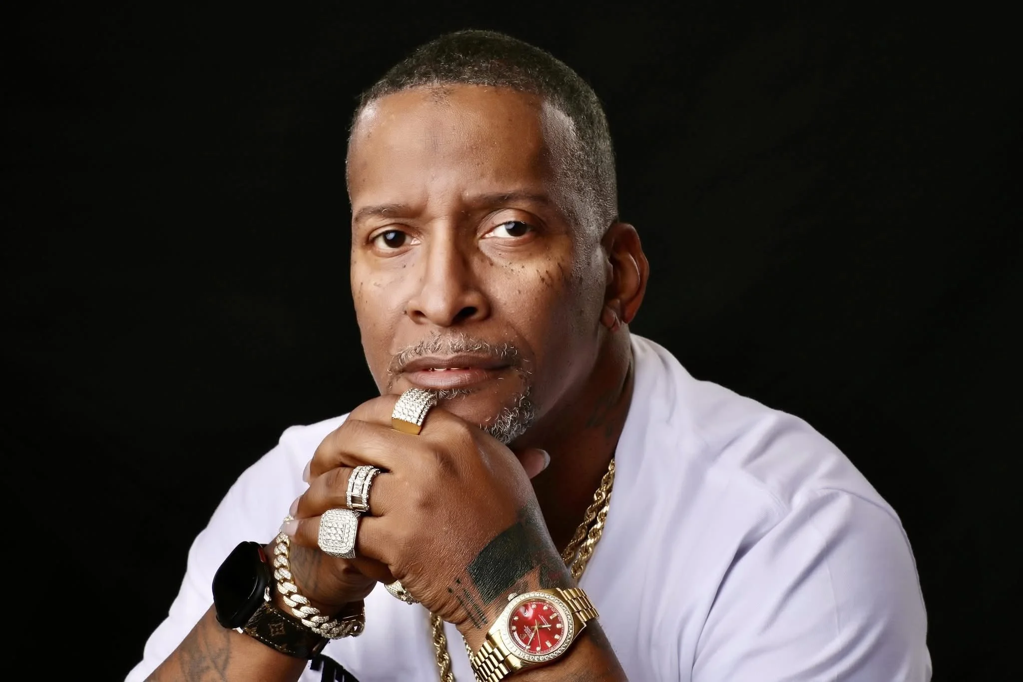 A man with short hair and a goatee wearing jewelry and a white shirt, resting his chin on his hand, against a black background.